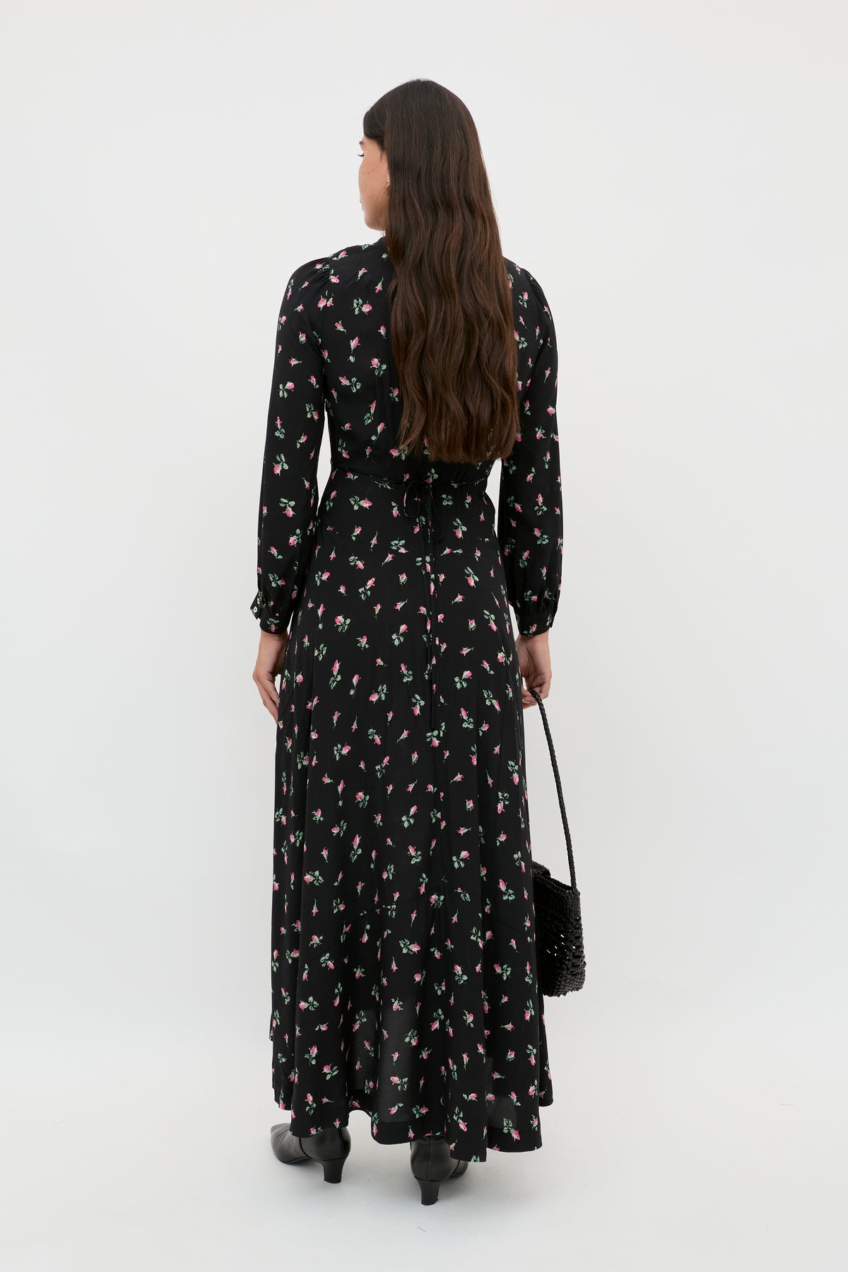 A woman with long brown hair stands facing away from the camera, holding a black woven bag and wearing the Avalyn - Sienna Bud Black midi dress with small floral prints and vintage cuffs, paired with black boots, against a white background.