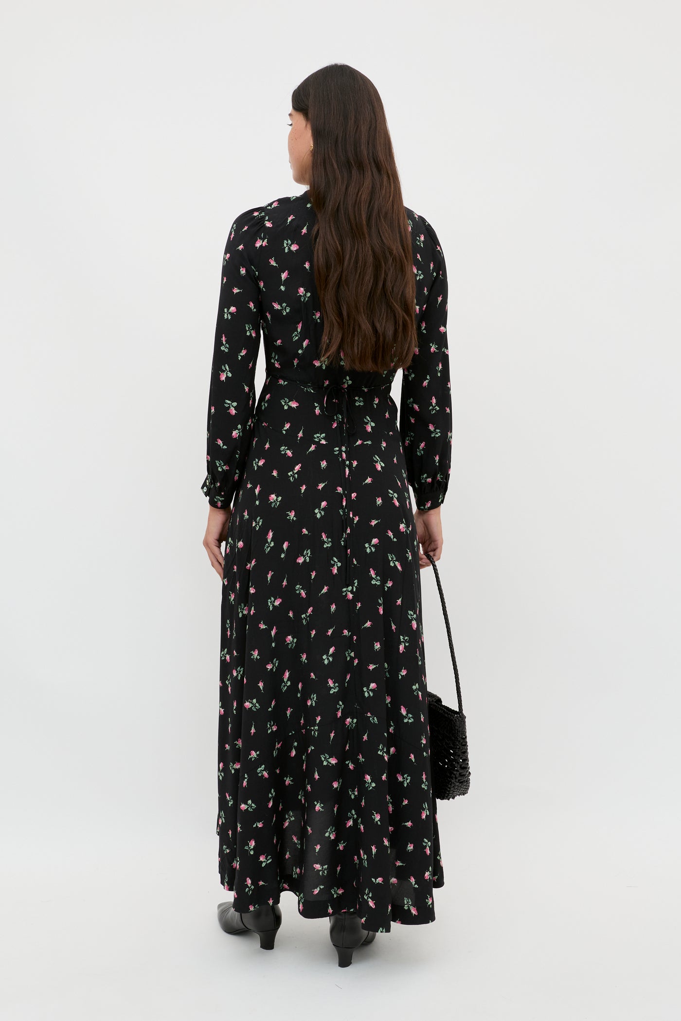 A woman with long brown hair stands facing away from the camera, holding a black woven bag and wearing the Avalyn - Sienna Bud Black midi dress with small floral prints and vintage cuffs, paired with black boots, against a white background.