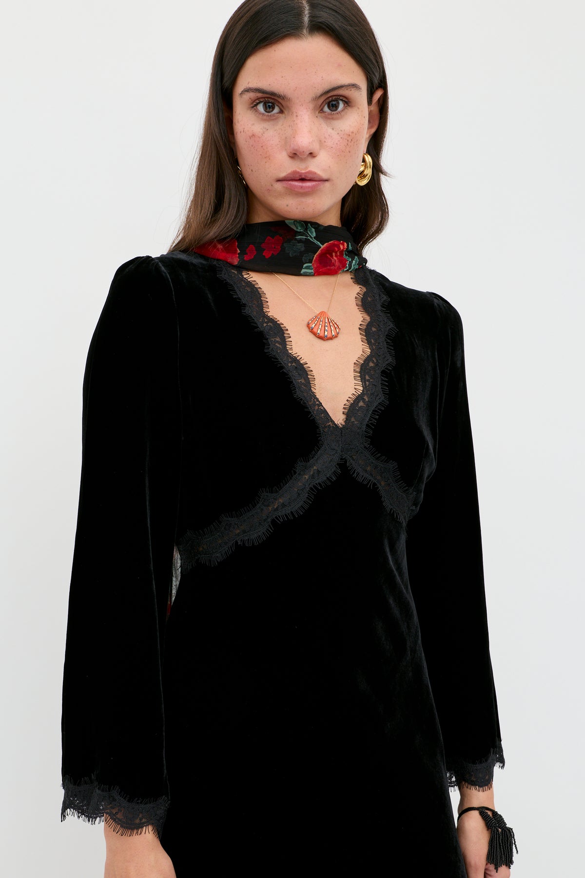 A woman with long brown hair and freckles wears the Alessi - Black velvet dress with lace details, paired with a red floral scarf, gold earrings, and a seashell pendant necklace, standing against a plain white background.