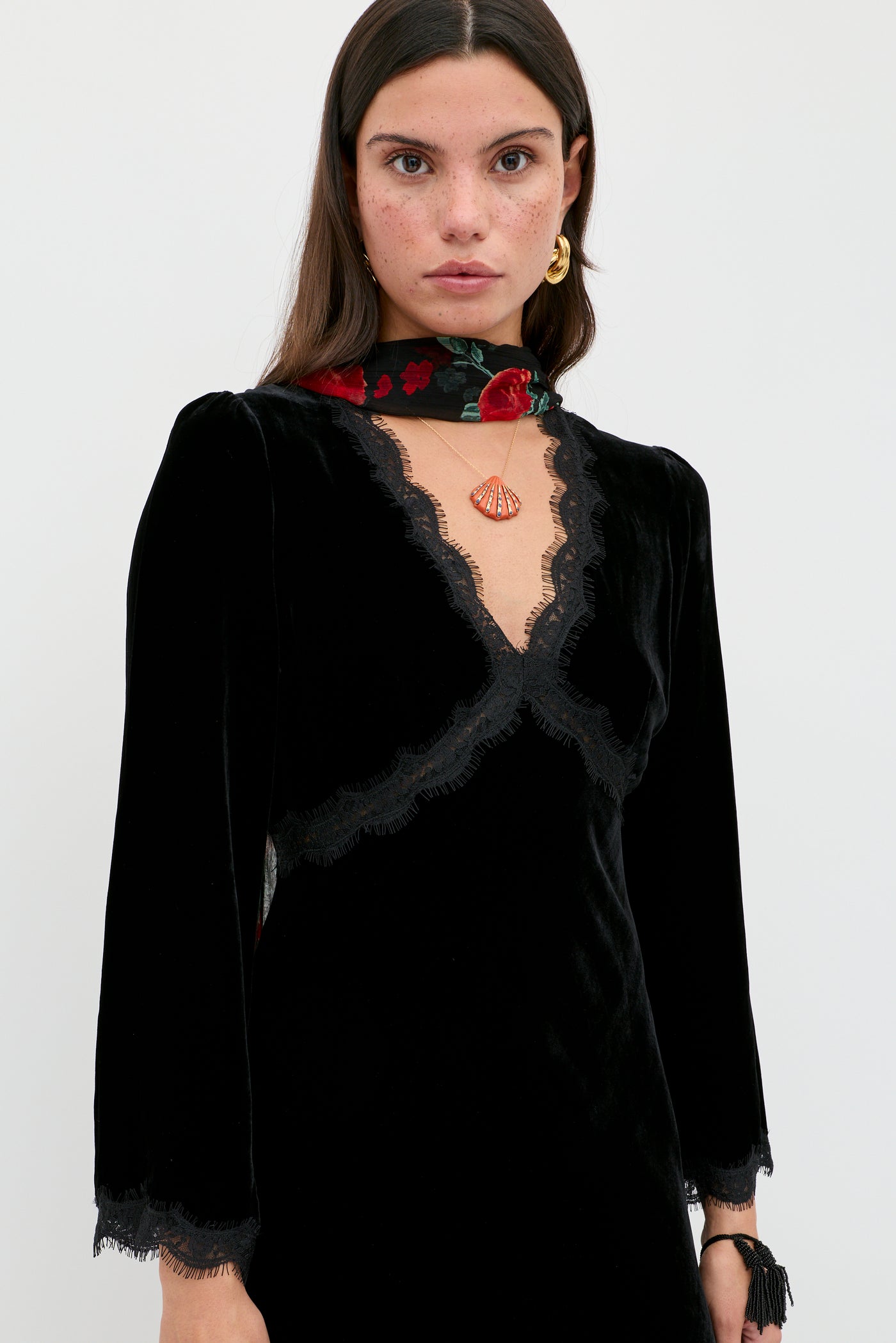 A woman with long brown hair and freckles wears the Alessi - Black velvet dress with lace details, paired with a red floral scarf, gold earrings, and a seashell pendant necklace, standing against a plain white background.