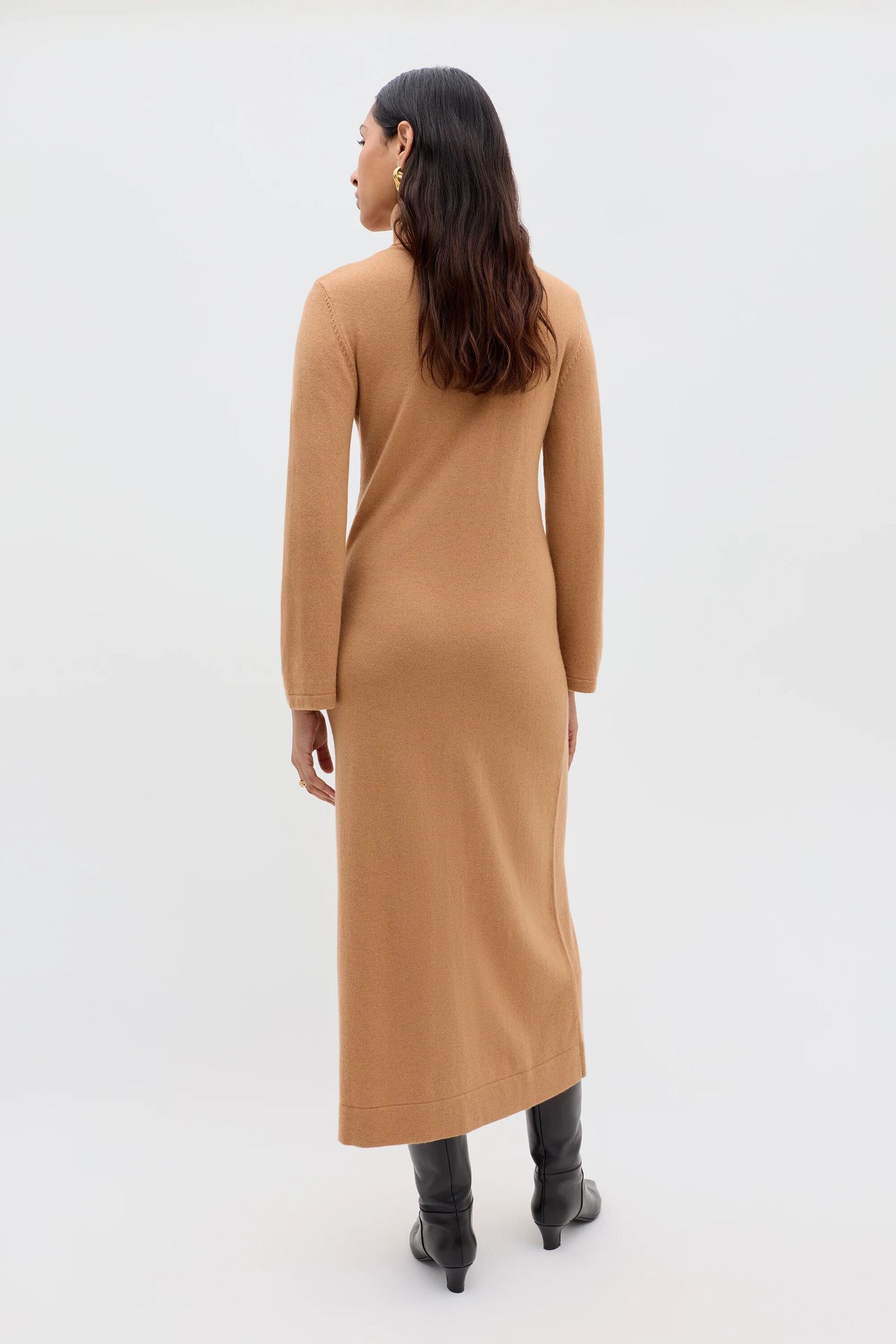 A person with long, wavy dark hair faces away in minimalist style, wearing the Elliette - Camel turtleneck knitted dress and black knee-high boots against a plain light background.