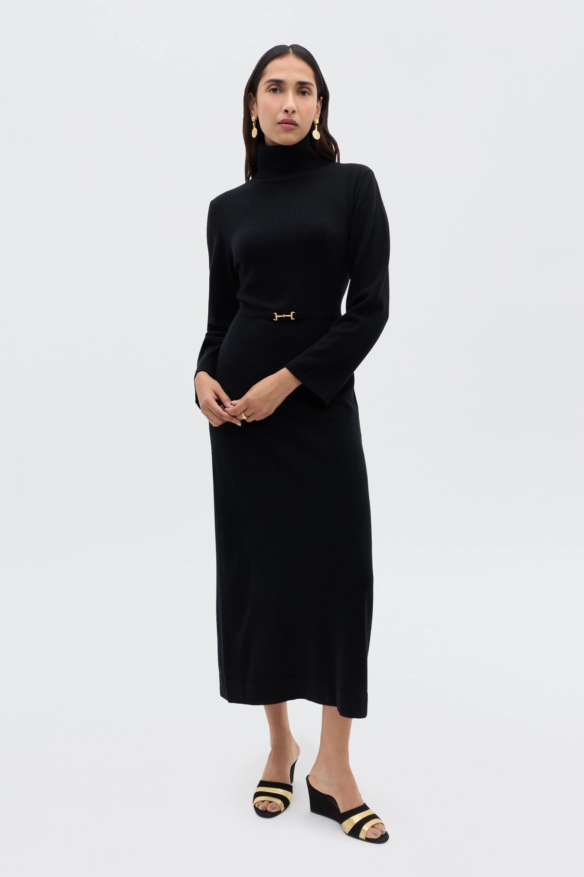 A woman with straight dark hair and a neutral expression poses against a light background, wearing the Elliette - Black long-sleeved turtleneck dress with gold waist detail, styled with black open-toe sandals featuring gold accents.