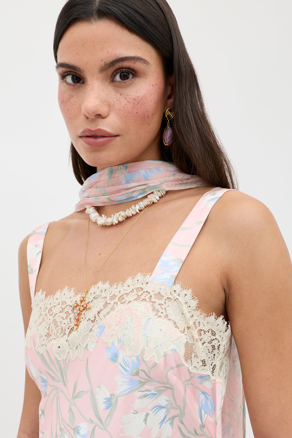 A woman with long dark hair wears the Rosamarie - Watercolour Floral Pink dress, styled with a sheer scarf, shell necklace, gold earrings, and a delicate pendant. She gazes at the camera with a neutral expression.