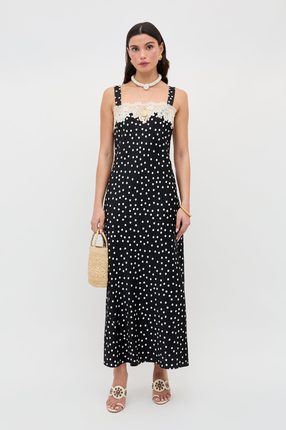 A woman stands against a plain white background wearing the Rosamarie - Scattered Dot Black dress with lace neckline, paired with beige sandals, a shell necklace, and holding a small woven handbag.