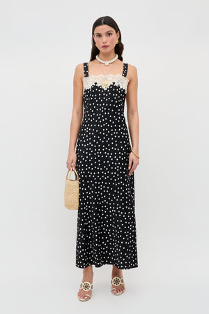 A woman stands against a plain white background wearing the Rosamarie - Scattered Dot Black dress with lace neckline, paired with beige sandals, a shell necklace, and holding a small woven handbag.
