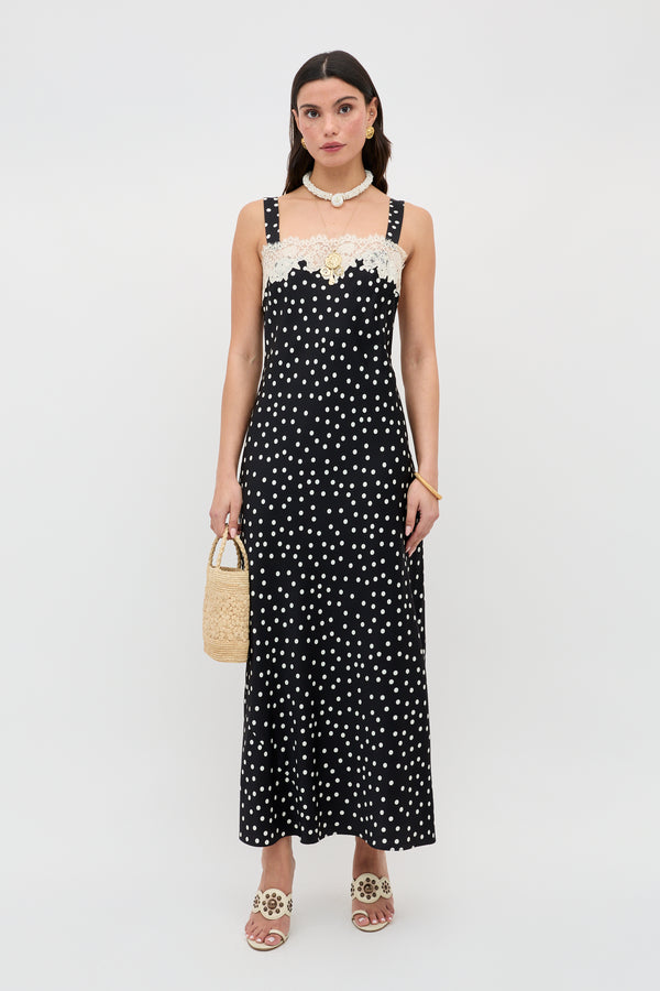 A woman stands against a plain white background wearing the Rosamarie - Scattered Dot Black dress with lace neckline, paired with beige sandals, a shell necklace, and holding a small woven handbag.