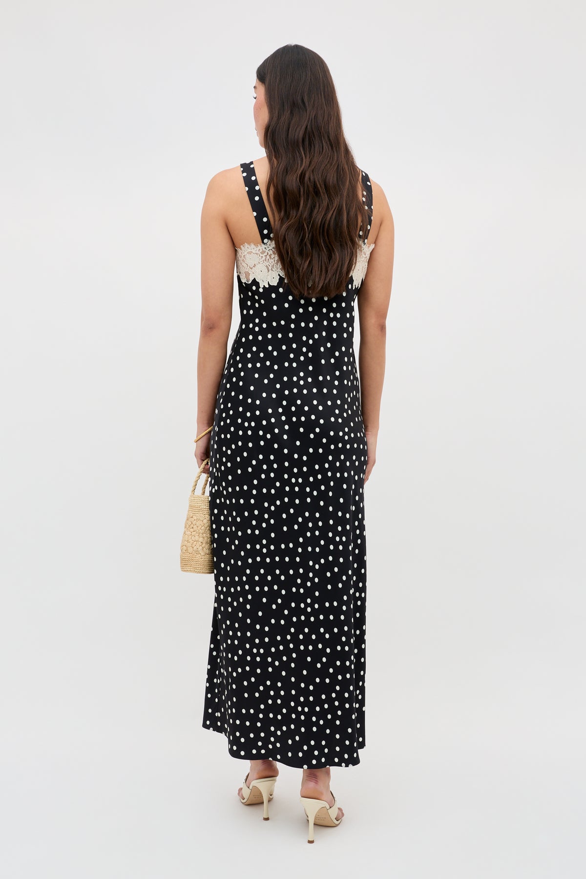 A woman with long, wavy brown hair stands facing away in the Rosamarie - Scattered Dot Black sleeveless maxi dress with white polka dots and lace trim, holding a woven handbag and wearing heeled sandals against a plain light background.