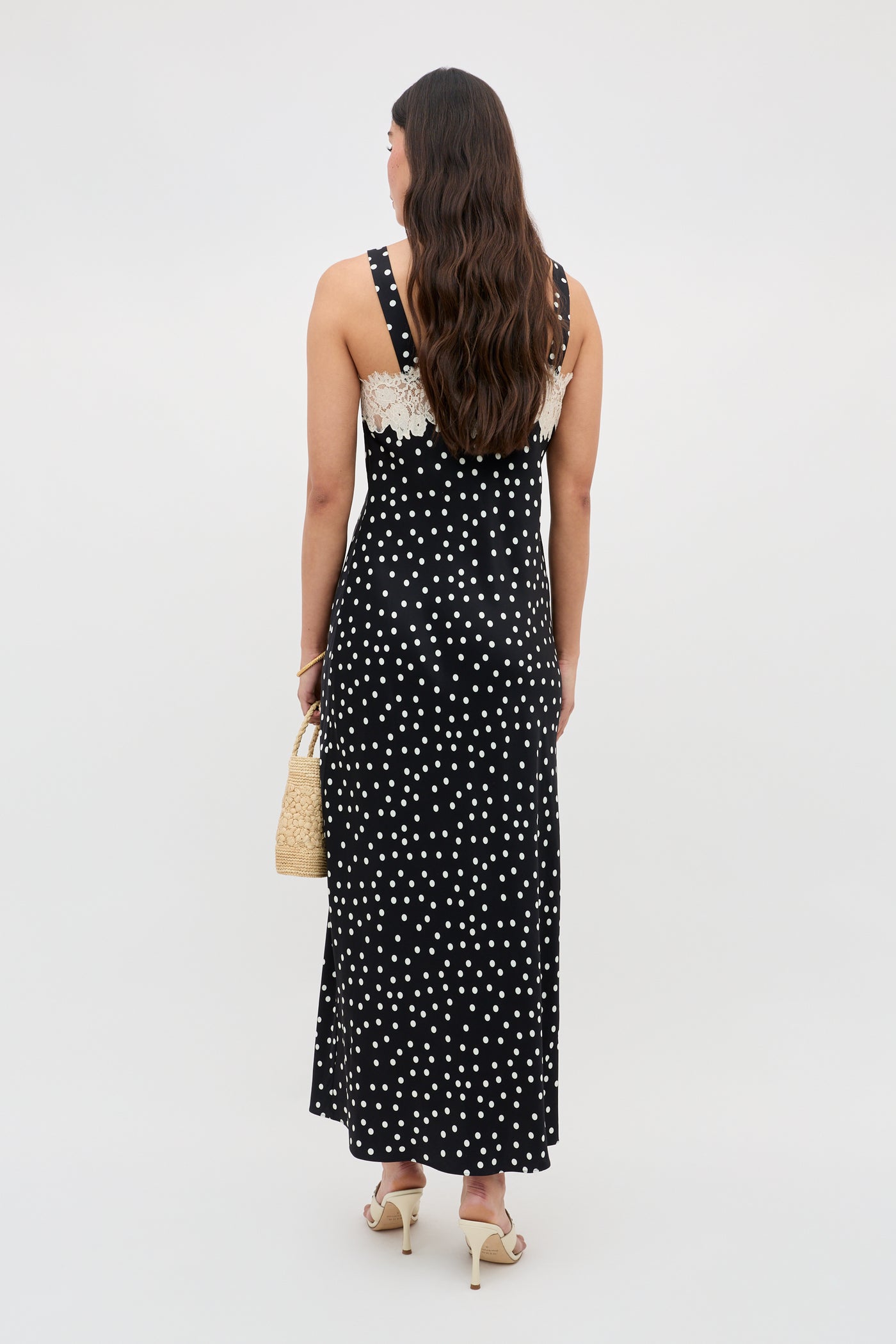 A woman with long, wavy brown hair stands facing away in the Rosamarie - Scattered Dot Black sleeveless maxi dress with white polka dots and lace trim, holding a woven handbag and wearing heeled sandals against a plain light background.