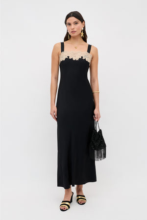 A woman with long dark hair and gold jewelry stands against a white background wearing the Rosamarie - Black sleeveless dress with beige lace at the neckline, holding a black beaded fringe bag.