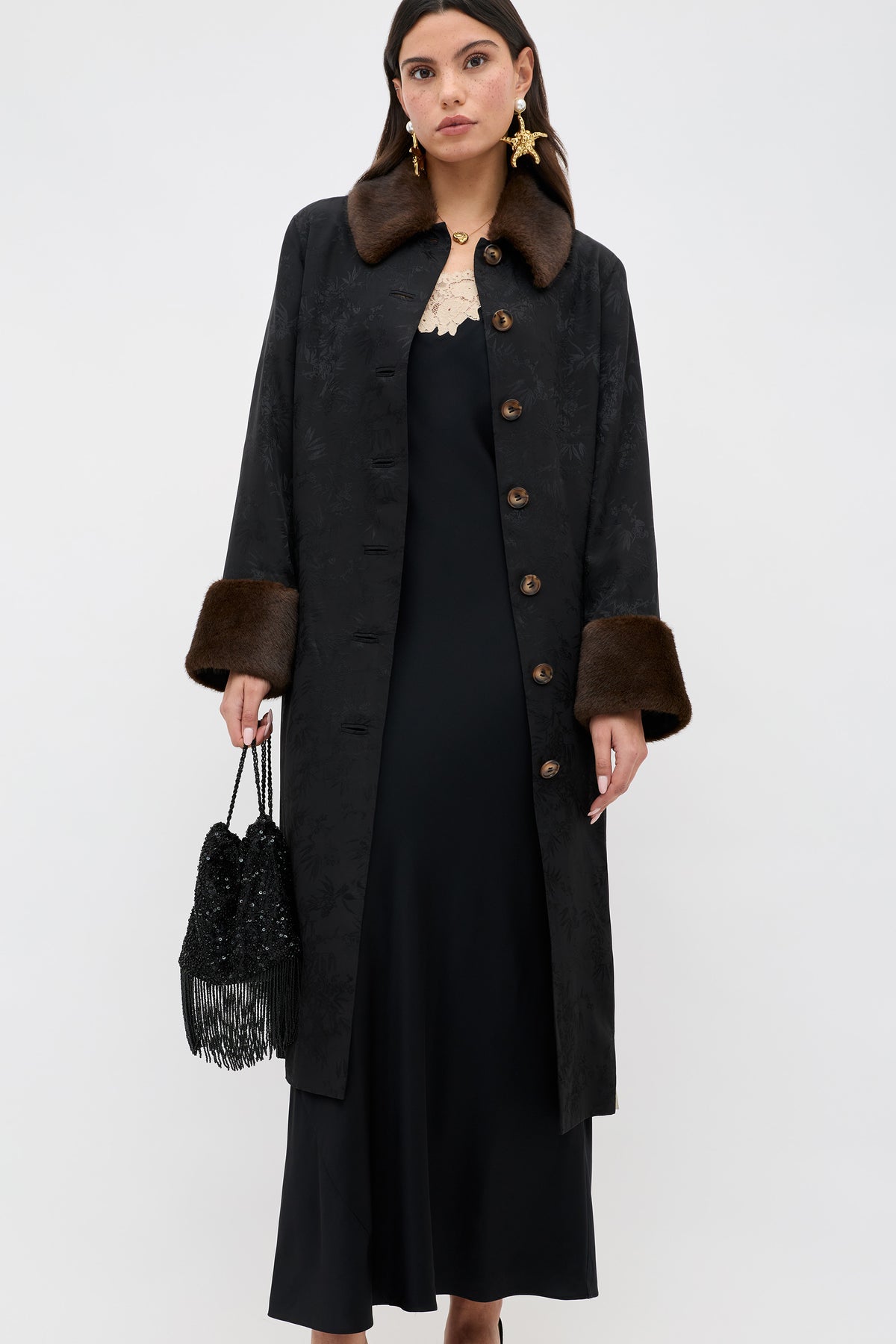 A woman wears the Rosamarie - Black long coat with brown fur collar and cuffs over a black dress, holding a black beaded fringe handbag and wearing statement earrings against a plain white background.