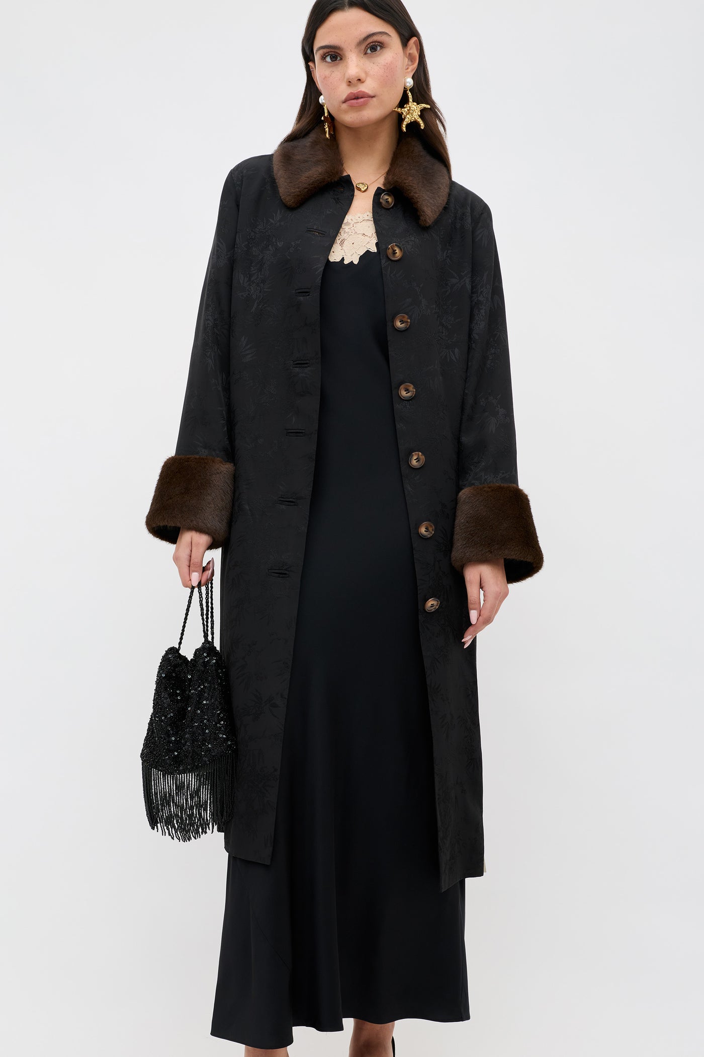 A woman wears the Rosamarie - Black long coat with brown fur collar and cuffs over a black dress, holding a black beaded fringe handbag and wearing statement earrings against a plain white background.