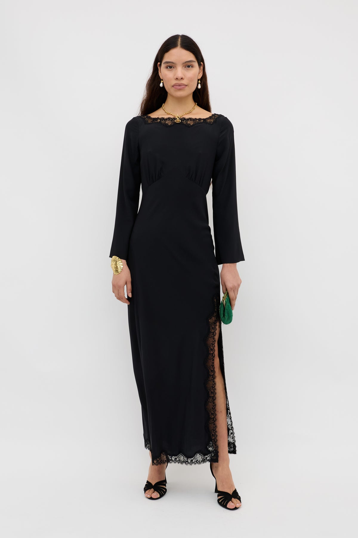 A woman stands against a white background wearing the Mirena - Black dress, a long black gown with lace trim and a high side slit. She pairs it with black heels, gold jewelry, and holds a small green bag.