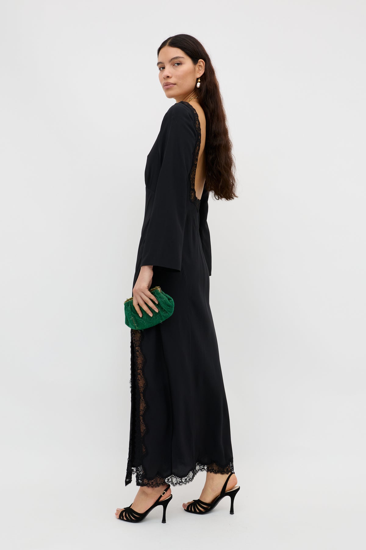 A woman stands sideways against a white background, wearing the Mirena - Black long-sleeved open-back dress with lace trim, black heeled sandals, gold earrings, and holding a small green clutch. She has long brown hair.
