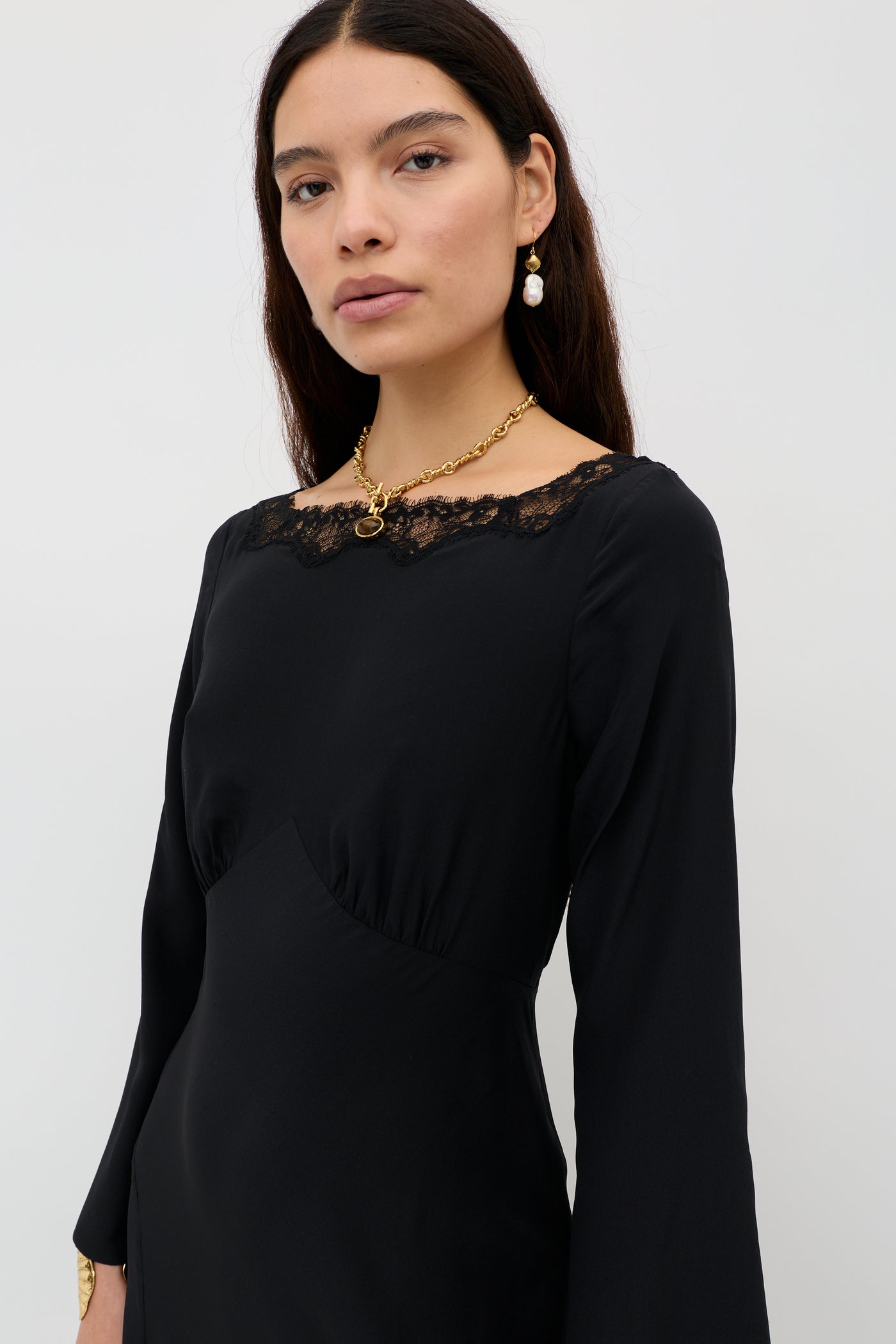 A woman with long dark hair wears the Mirena - Black dress with lace neckline trim, paired with a gold chain necklace and gold drop earrings, standing against a plain light background.