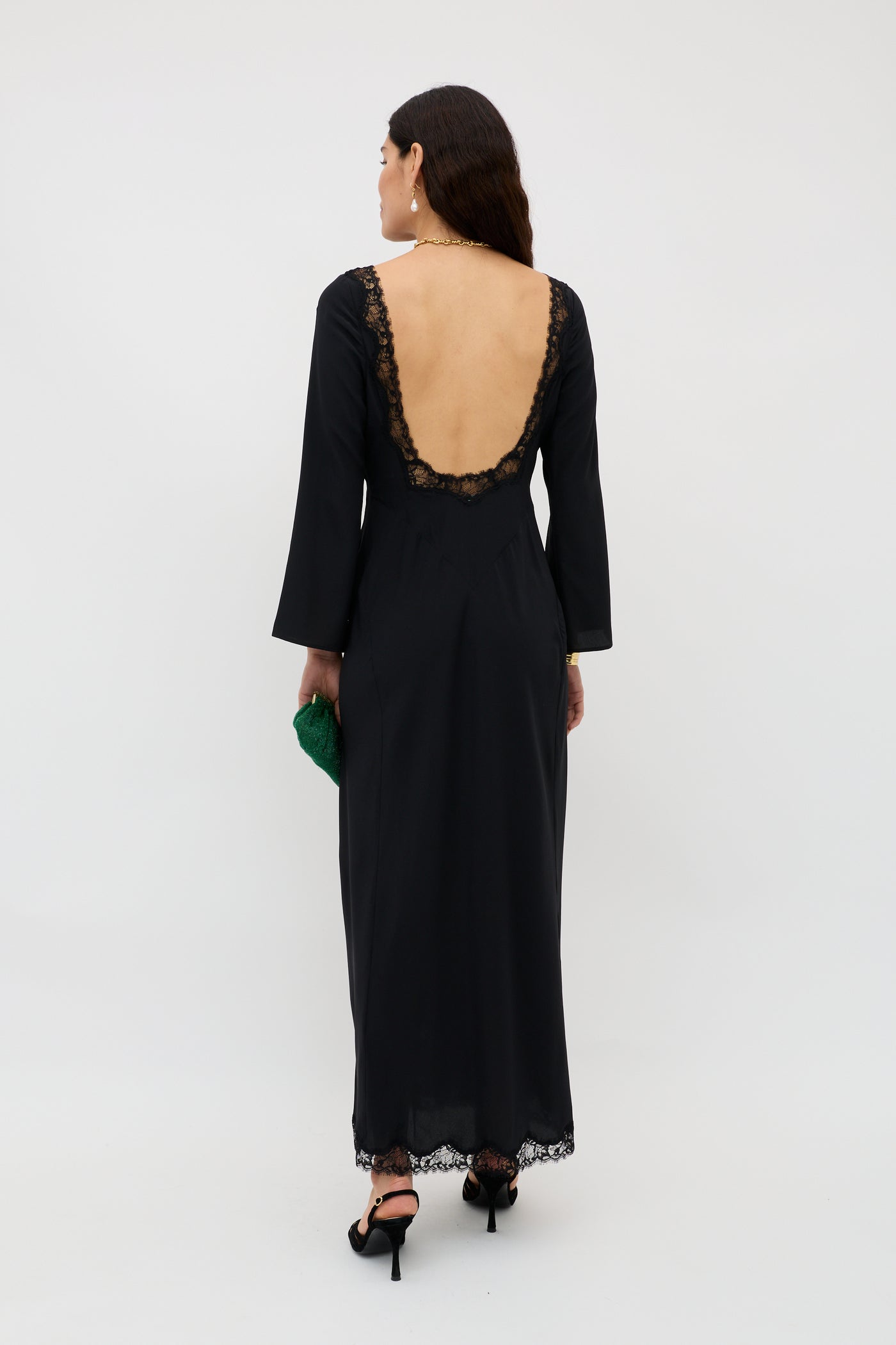 A woman wearing the Mirena - Black dress with lace trim and an open back stands facing away, holding a green clutch. She has long brown hair, black heels, and earrings. The background is plain and light-colored.