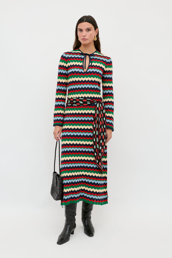 A woman is wearing the Lucila - Retro Chevron Blue long-sleeved dress, featuring a multicolored wavy chevron pattern and a keyhole neckline, styled with a patterned belt, black boots, and a black textured handbag, standing against a plain white background.