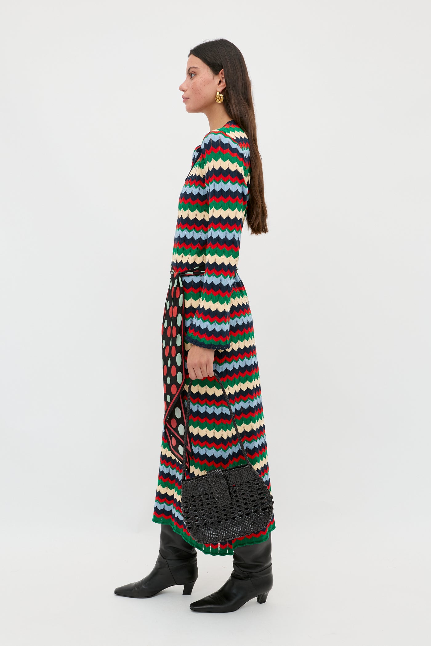 A woman wearing the Lucila - Retro Chevron Blue dress, featuring long sleeves and a colorful wavy pattern, paired with black boots and a black woven handbag, stands sideways against a plain white background.