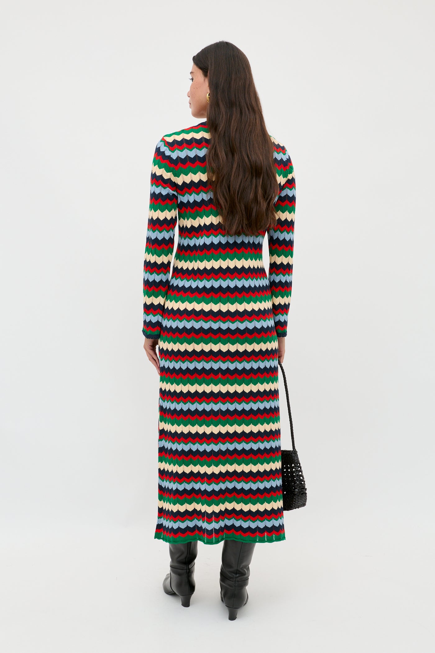 A person with long brown hair, wearing the Lucila - Retro Chevron Blue dress and black boots, stands facing away and holds a black woven bag.