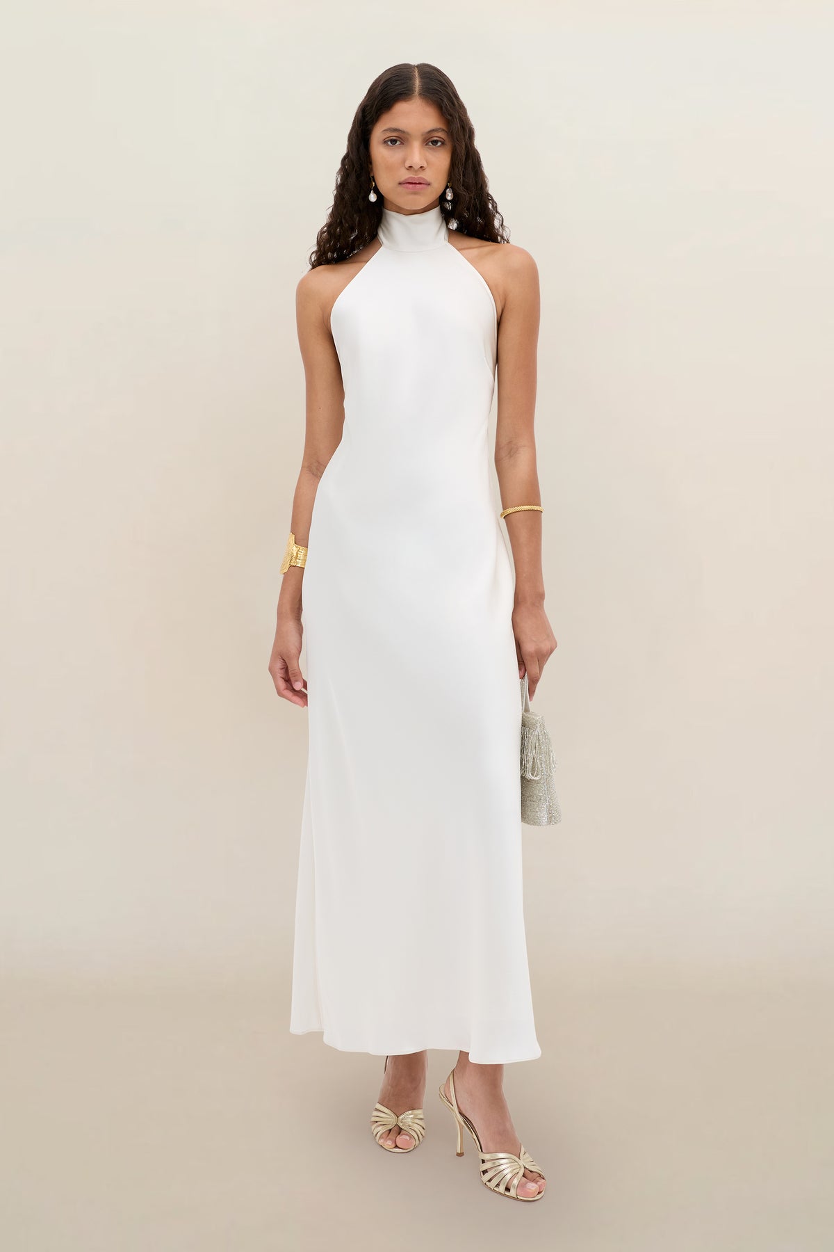 A woman wearing the Arista - Pearl sleeveless, high-neck, floor-length white dress with open shoulders, paired with gold strappy heels, gold jewelry, and a small handbag, stands against a plain beige background.