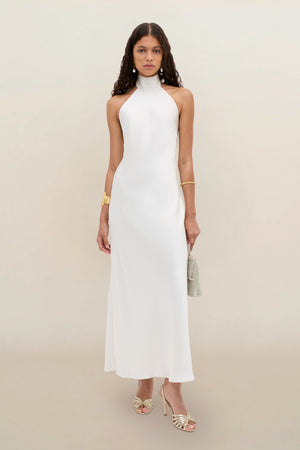 A woman wearing the Arista - Pearl sleeveless, high-neck, floor-length white dress with open shoulders, paired with gold strappy heels, gold jewelry, and a small handbag, stands against a plain beige background.