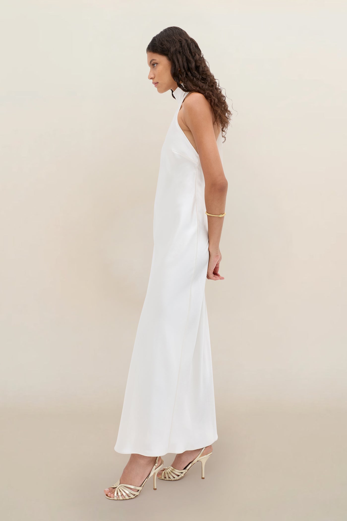 A woman in profile models the Arista - Pearl: a sleeveless, floor-length white dress, styled with gold heels and a gold bracelet. Her long wavy hair and a simple light background complete the look.