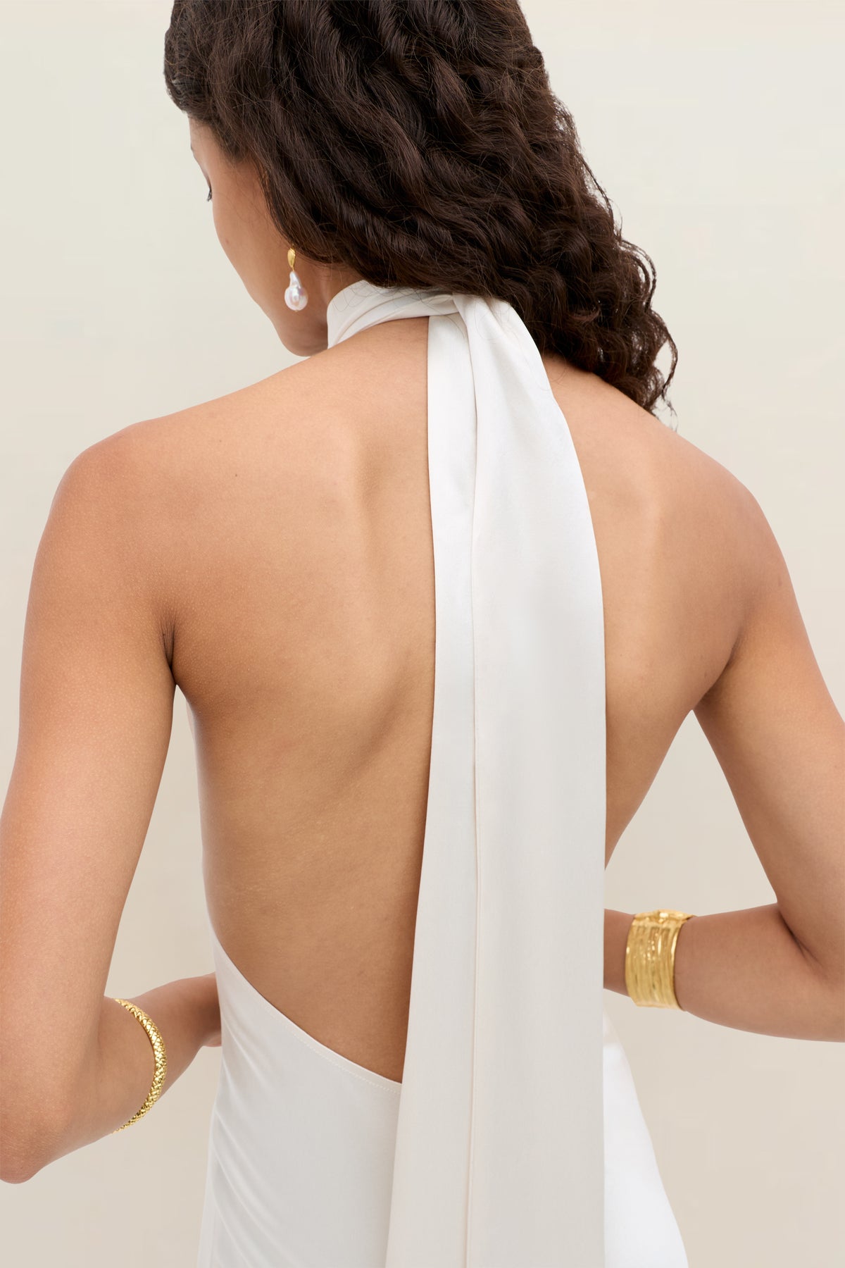 A woman with wavy dark hair wears the Arista – Pearl backless white halter dress featuring a long scarf detail. She accessorizes with gold bangles, a ring, and pearl drop earrings against a plain light-colored background.