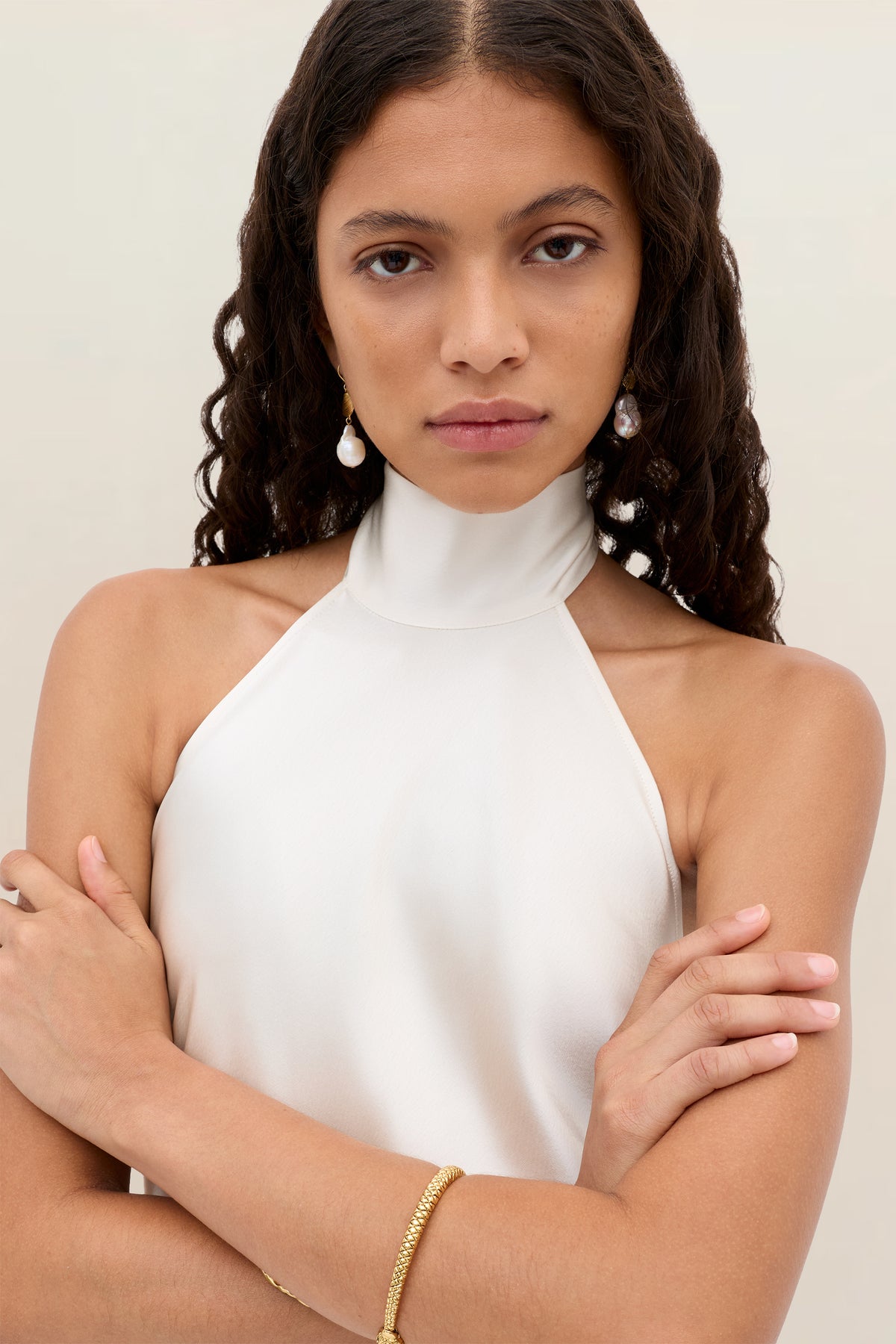 A woman with long curly hair stands with arms crossed, wearing a sleeveless white halter top, gold bracelet, and Arista – Pearl earrings against a plain light background.