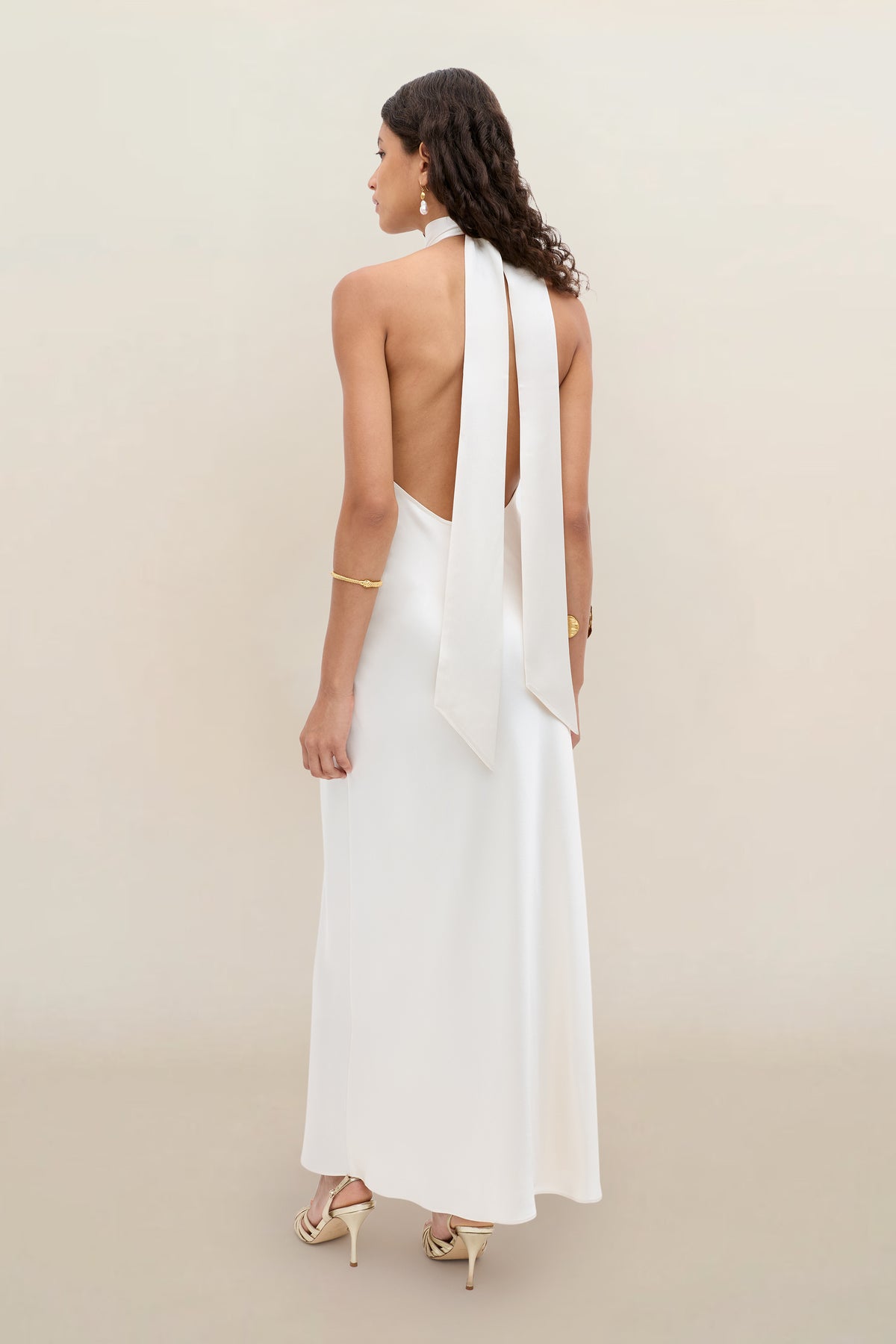 A woman with wavy dark hair models the Arista - Pearl, an elegant white halter dress with an open back and long ties, paired with gold jewelry and heeled sandals against a neutral beige background.