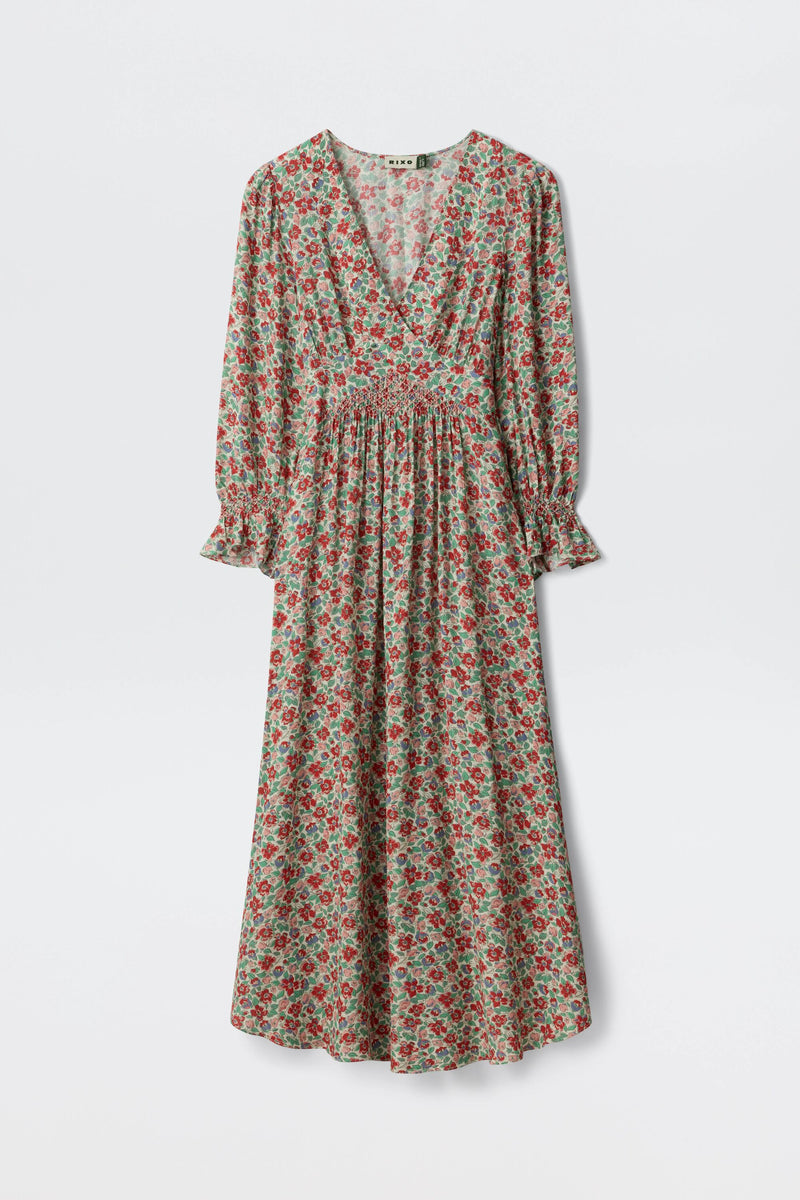 The Mireta - Jeannie Bloom Ivory is a long-sleeved, ankle-length dress with a red and green floral pattern, V-neckline, gathered waist, and slightly flared cuffs, displayed against a plain white background.