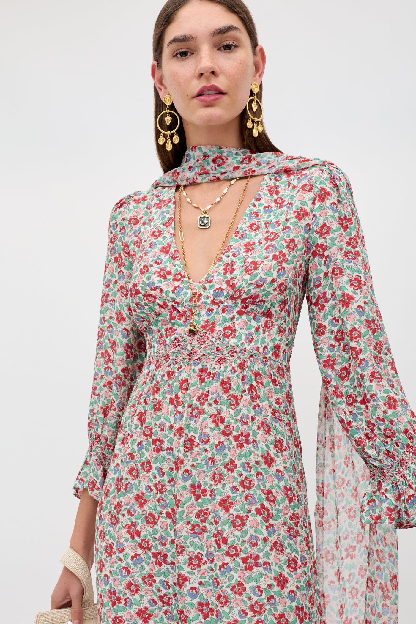 A woman models the Mireta - Jeannie Bloom Ivory, a long-sleeved floral dress with a deep V-neck, accessorized with gold earrings, layered necklaces, and a beige bag. She gazes confidently at the camera against a plain background.