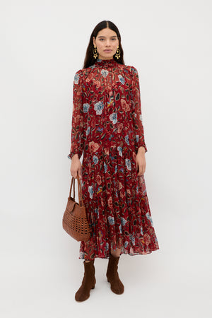 A woman wearing the Adalind - Butterfly Floral Burgundy dress with brown boots, gold earrings, and a brown woven handbag stands against a plain white background.