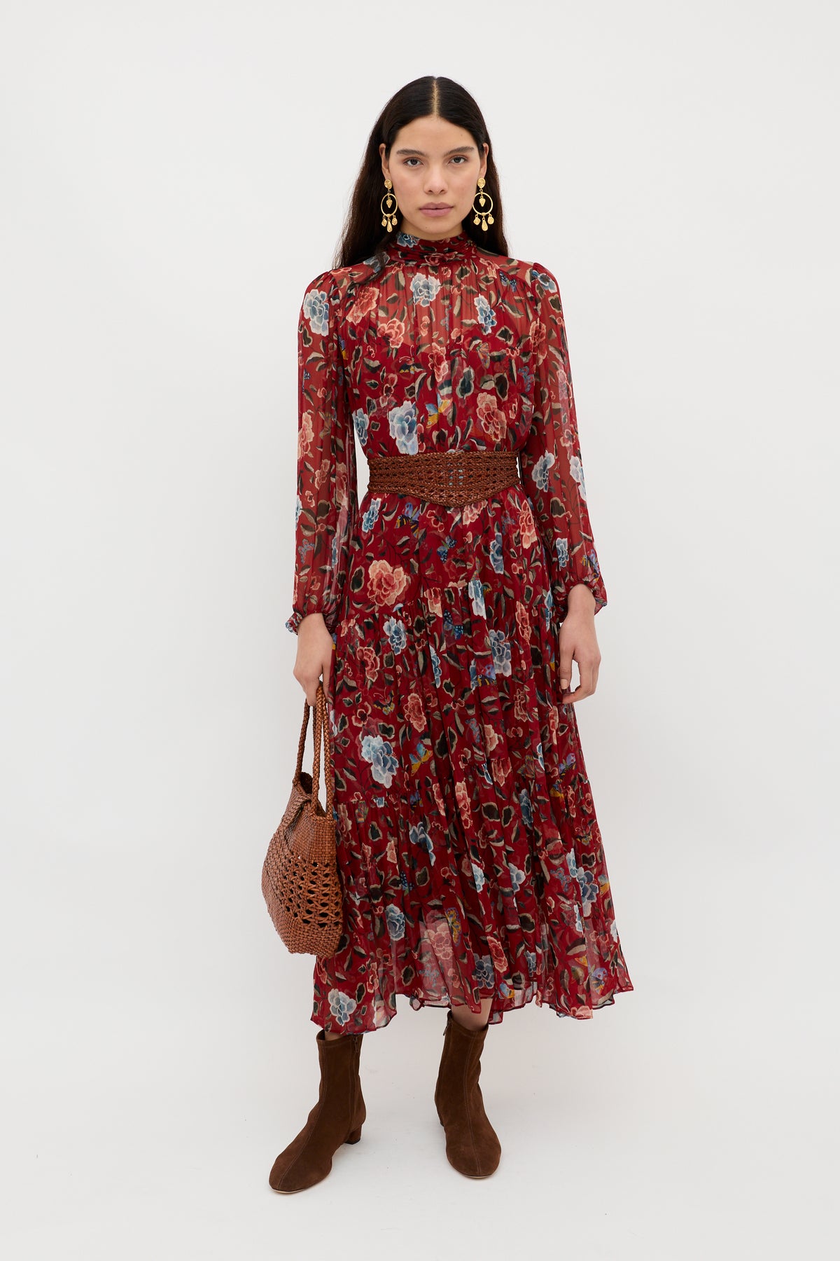 A woman wearing the Adalind - Butterfly Floral Burgundy chiffon dress with sheer sleeves, brown boots, gold earrings, and a brown woven handbag stands against a plain white background.