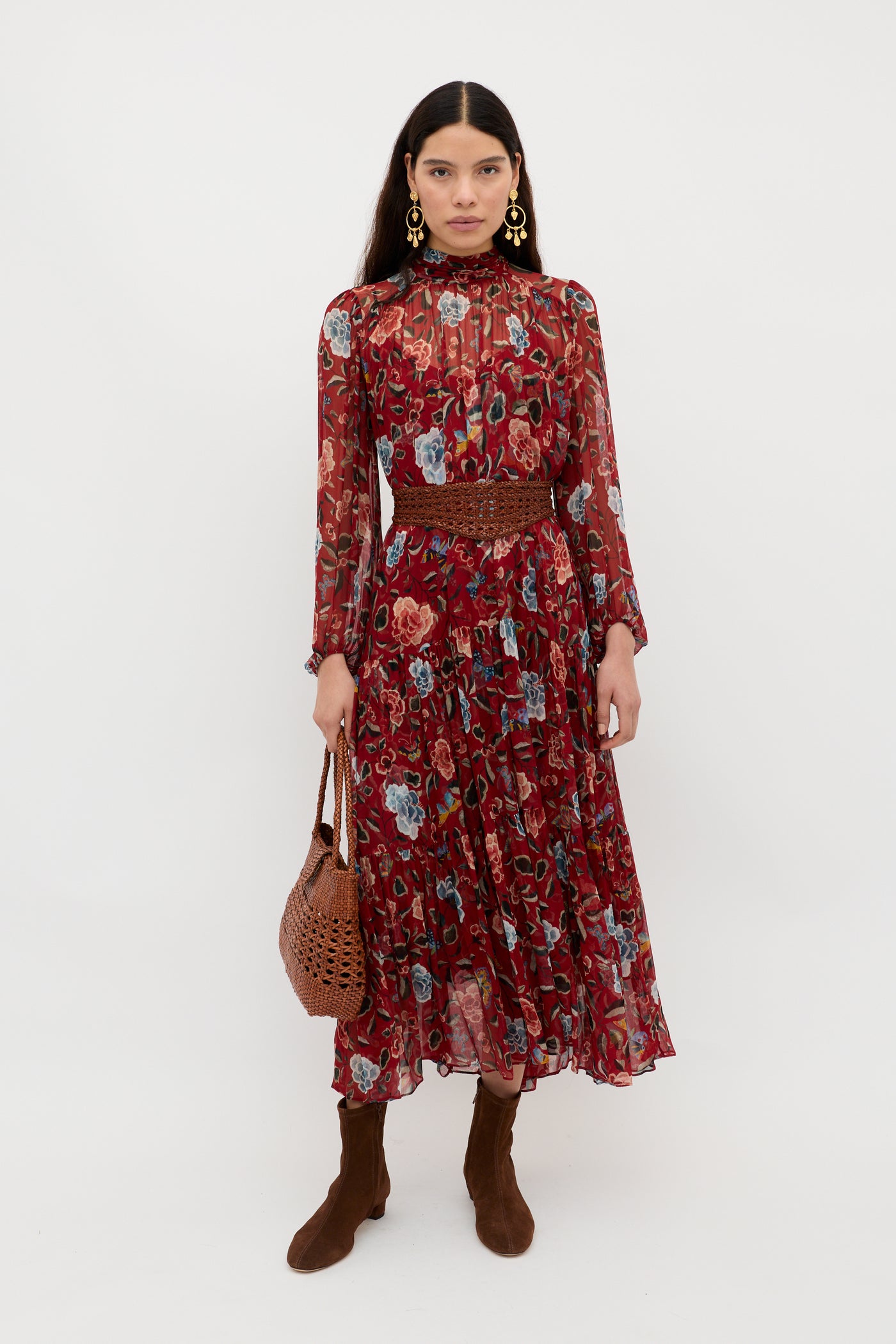 A woman wearing the Adalind - Butterfly Floral Burgundy chiffon dress with sheer sleeves, brown boots, gold earrings, and a brown woven handbag stands against a plain white background.