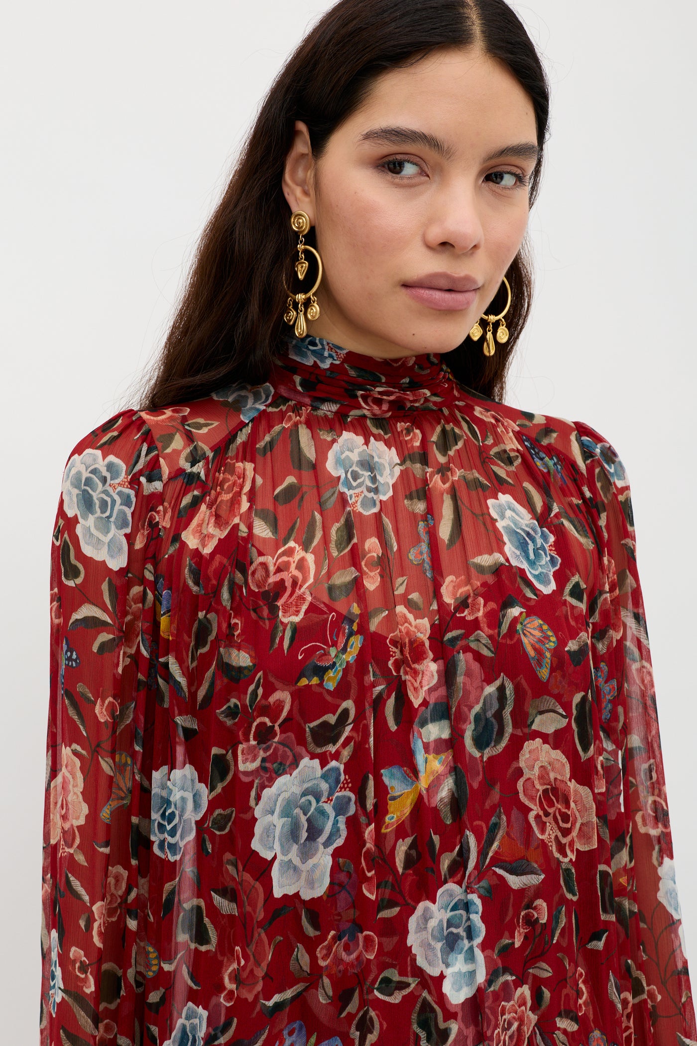 A woman with long dark hair wears gold earrings and the Adalind - Butterfly Floral Burgundy dress, featuring a high neck and vibrant floral print. She stands against a light background, gazing just past the camera.