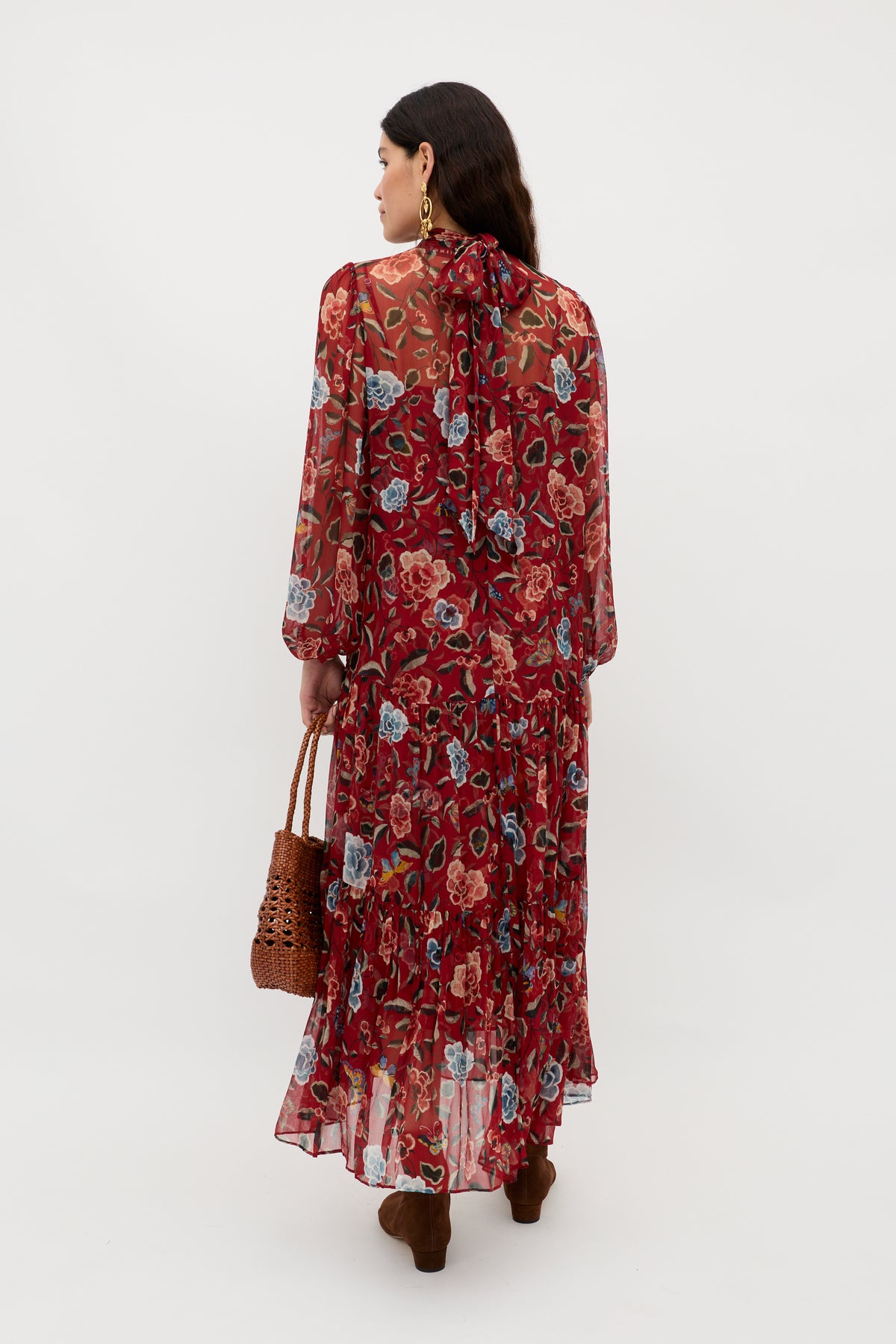 A woman with long dark hair, wearing gold hoop earrings and the Adalind - Butterfly Floral Burgundy dress, brown boots, and a woven handbag, stands facing away against a white background—an elegant eveningwear look.