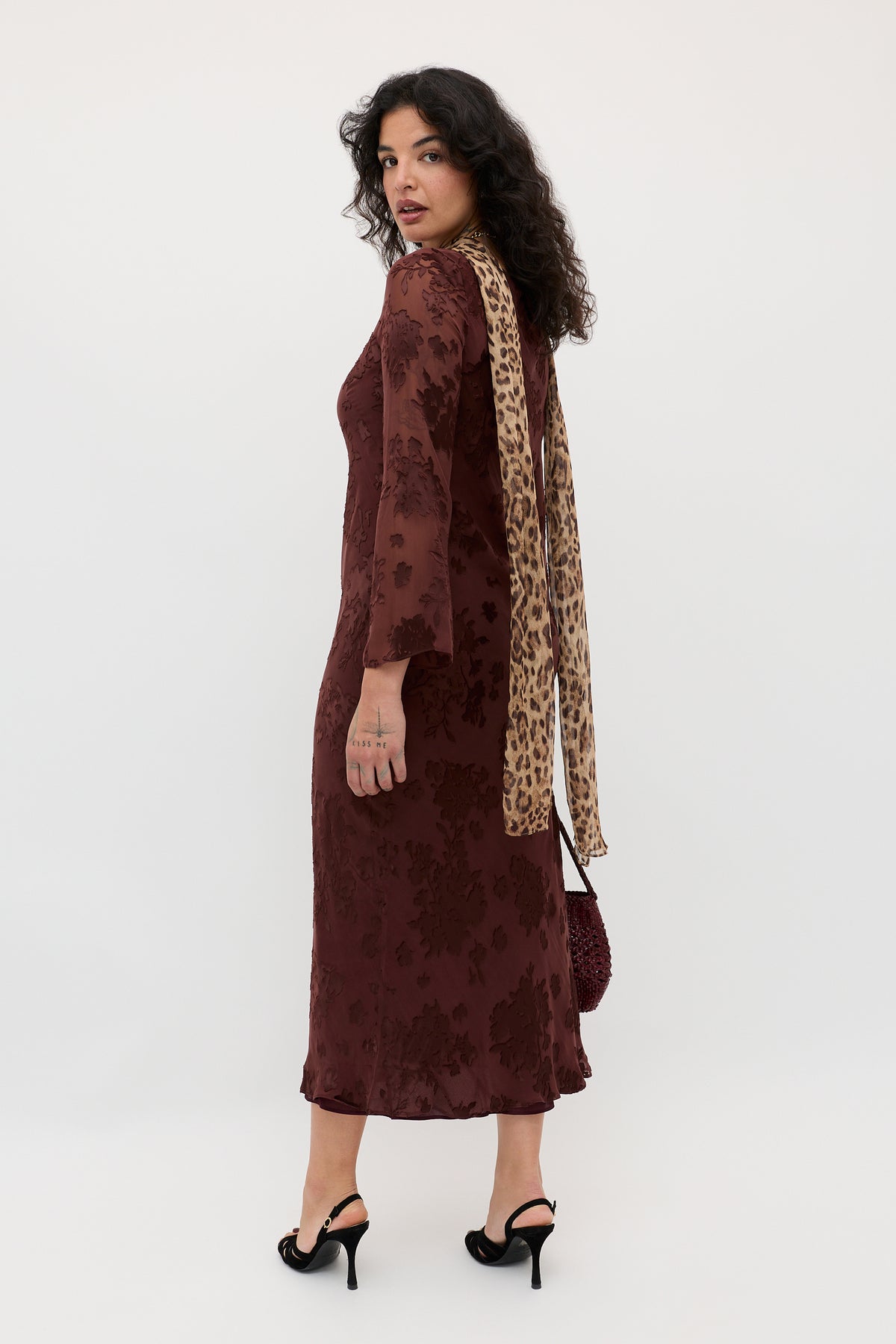 Wearing the Noraya - Bouquet Devoré Brown dress, a woman with curly dark hair stands in black heels, facing slightly away and looking back over her shoulder. She completes the look with a beige leopard-print scarf.