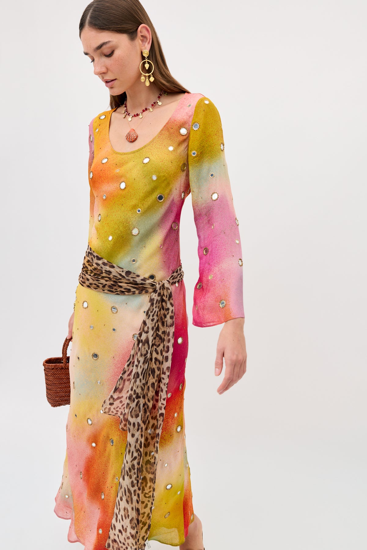 Dressed in the Noraya - Jessica Alexandra Multi, a vibrant tie-dye long-sleeved dress with sparkling sequin accents and a leopard print sash, this woman accessorizes with statement earrings, a necklace, and carries a brown woven handbag as she looks down against a plain background.