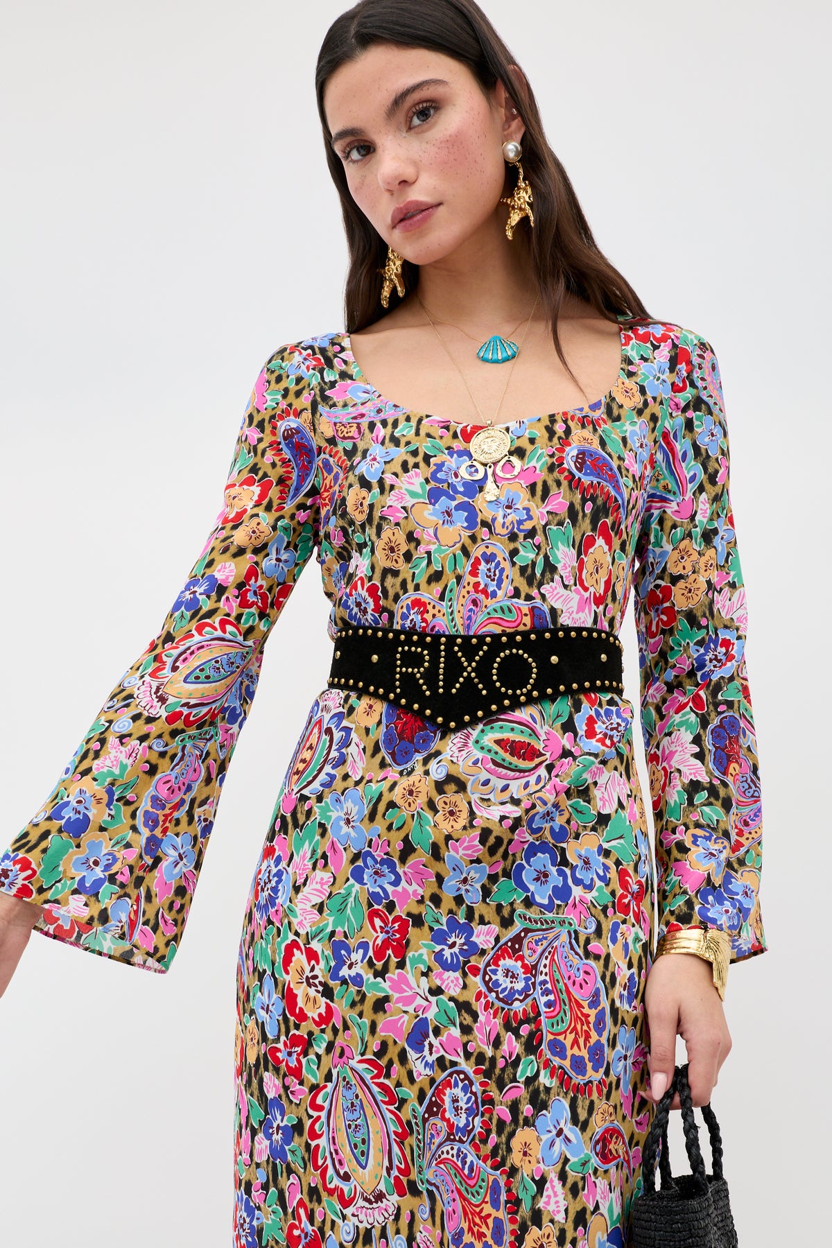 A woman in a colorful, long-sleeved floral and paisley print dress with a black studded belt, gold jewelry, Zeina - Gold clip-on earrings, and a turquoise necklace holds a textured black handbag against a plain background.