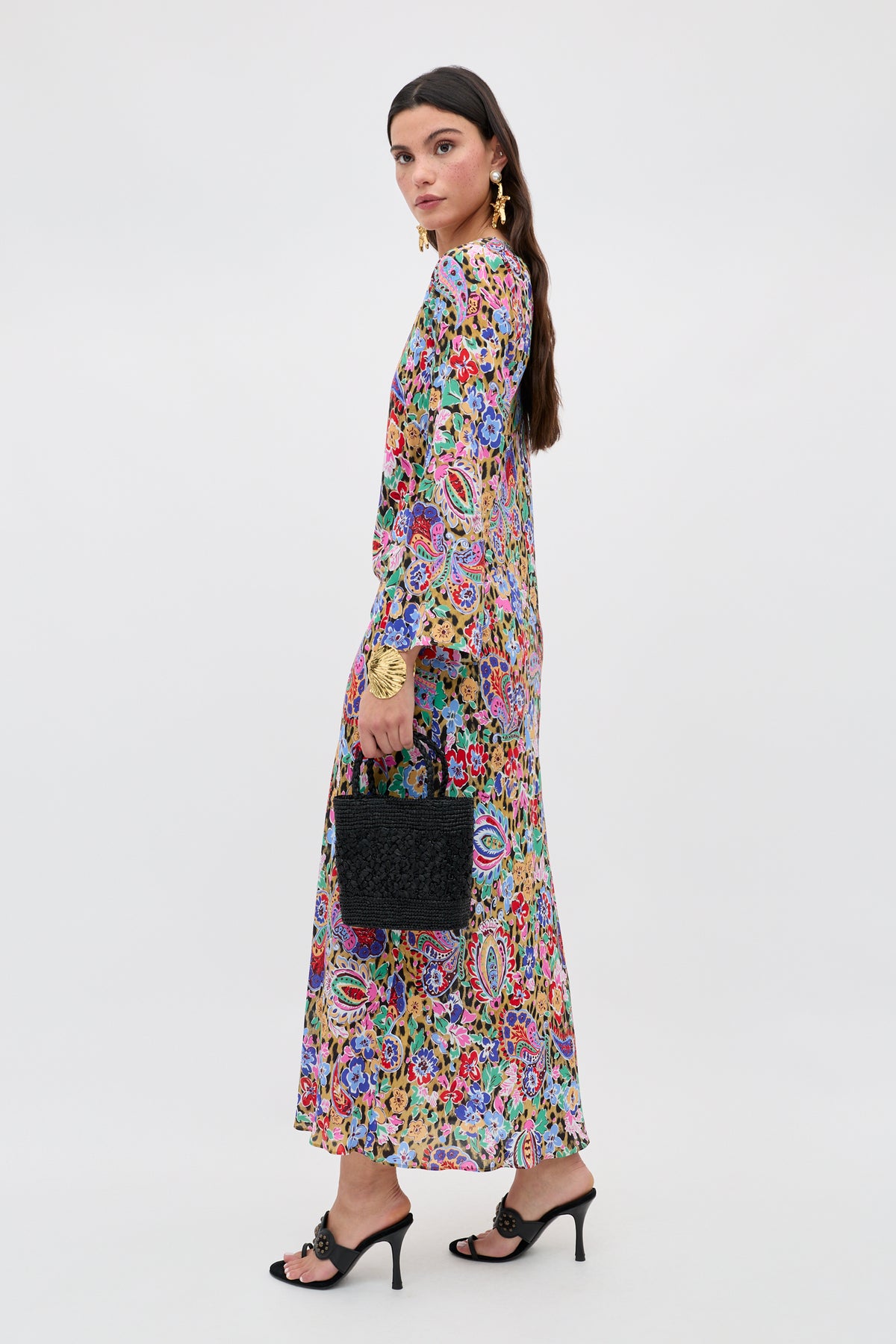 A woman stands sideways in the Noraya - Cara Floral Leopard Brown dress, holding a black textured handbag. She wears black heeled sandals and gold statement earrings, and her long brown hair is styled straight. The background is plain and light.