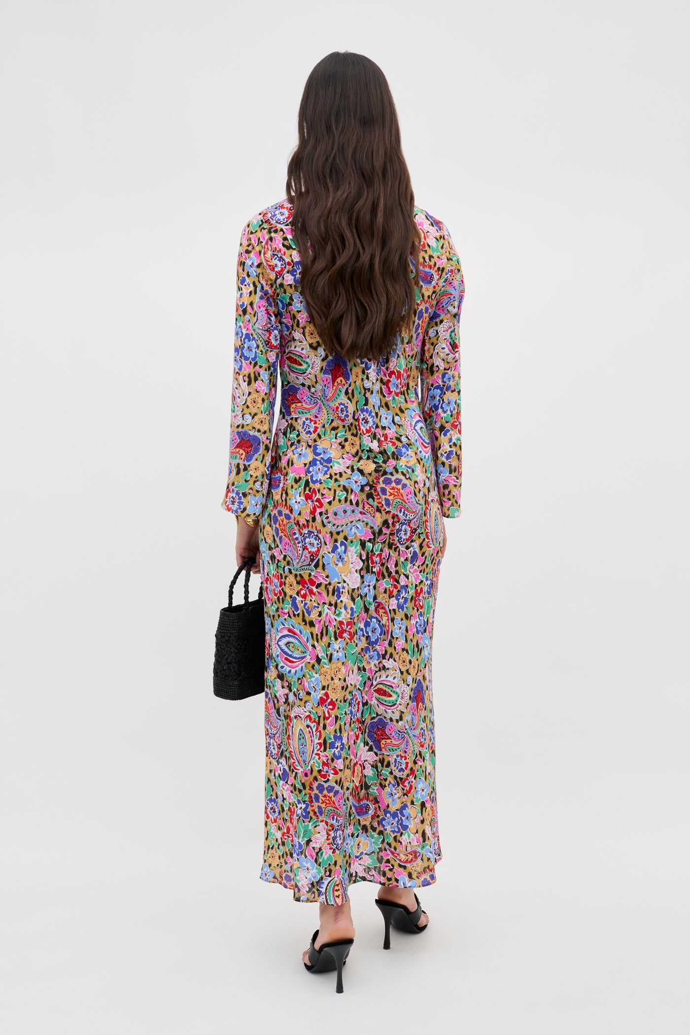 A person with long wavy hair, dressed in the Noraya - Cara Floral Leopard Brown maxi dress and black heeled sandals, stands facing away and holds a small black handbag against a plain light background.