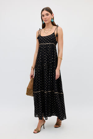 A woman wearing the Kamilah - Black sleeveless, ankle-length dress with gold polka dots, styled with strappy heels, gold jewelry, blue earrings, and a brown textured handbag, stands against a plain light background.