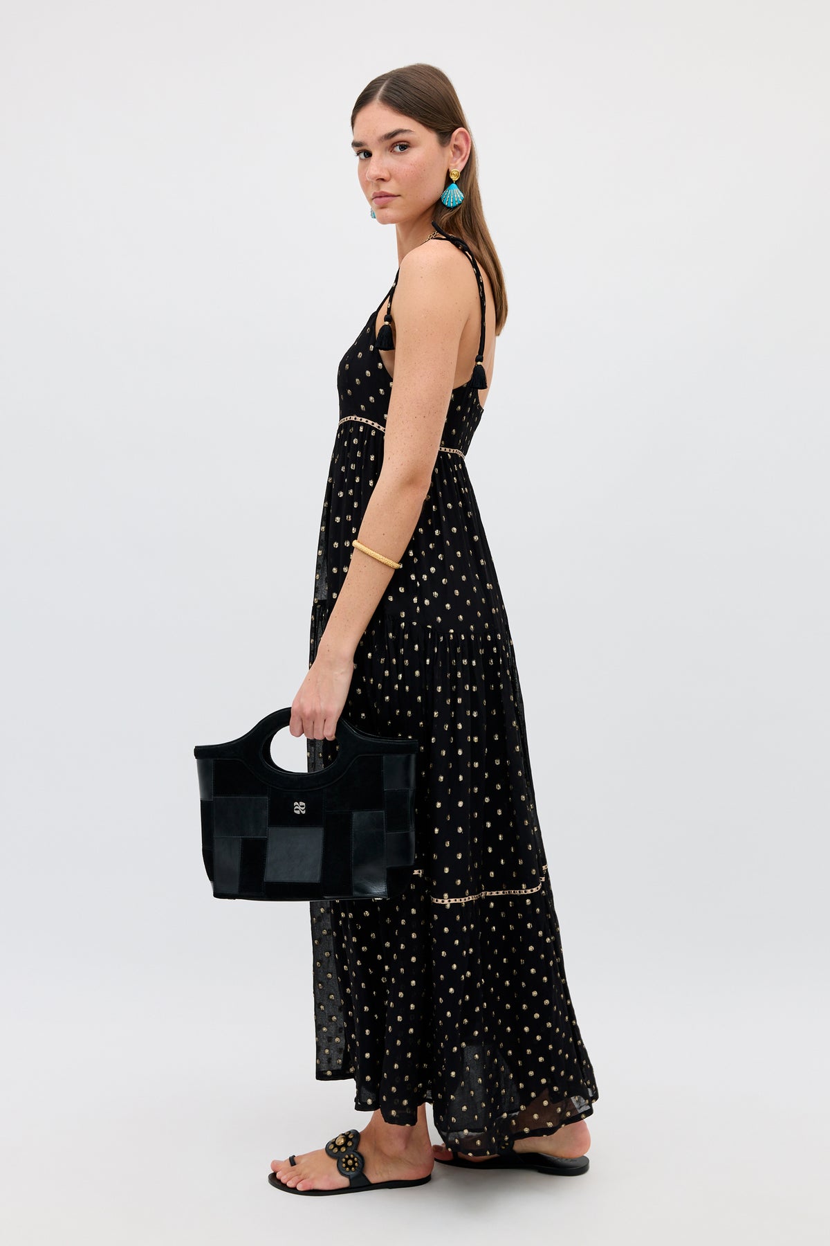 A woman in the “Kamilah - Black” sleeveless maxi dress with white polka dots, accessorized with a black handbag, black sandals, a gold bracelet, and turquoise earrings, stands sideways against a plain white background.