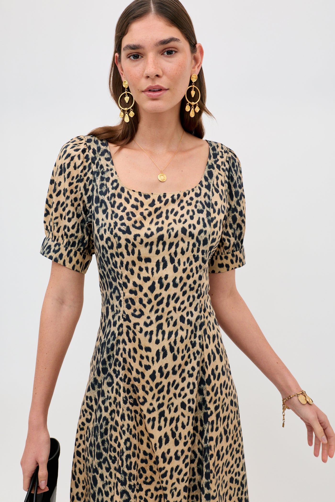 A woman stands against a white background, wearing the Kathleen - Nina Leopard Brown dress, paired with gold drop earrings, a gold necklace, a bracelet, and holding a small black bag.