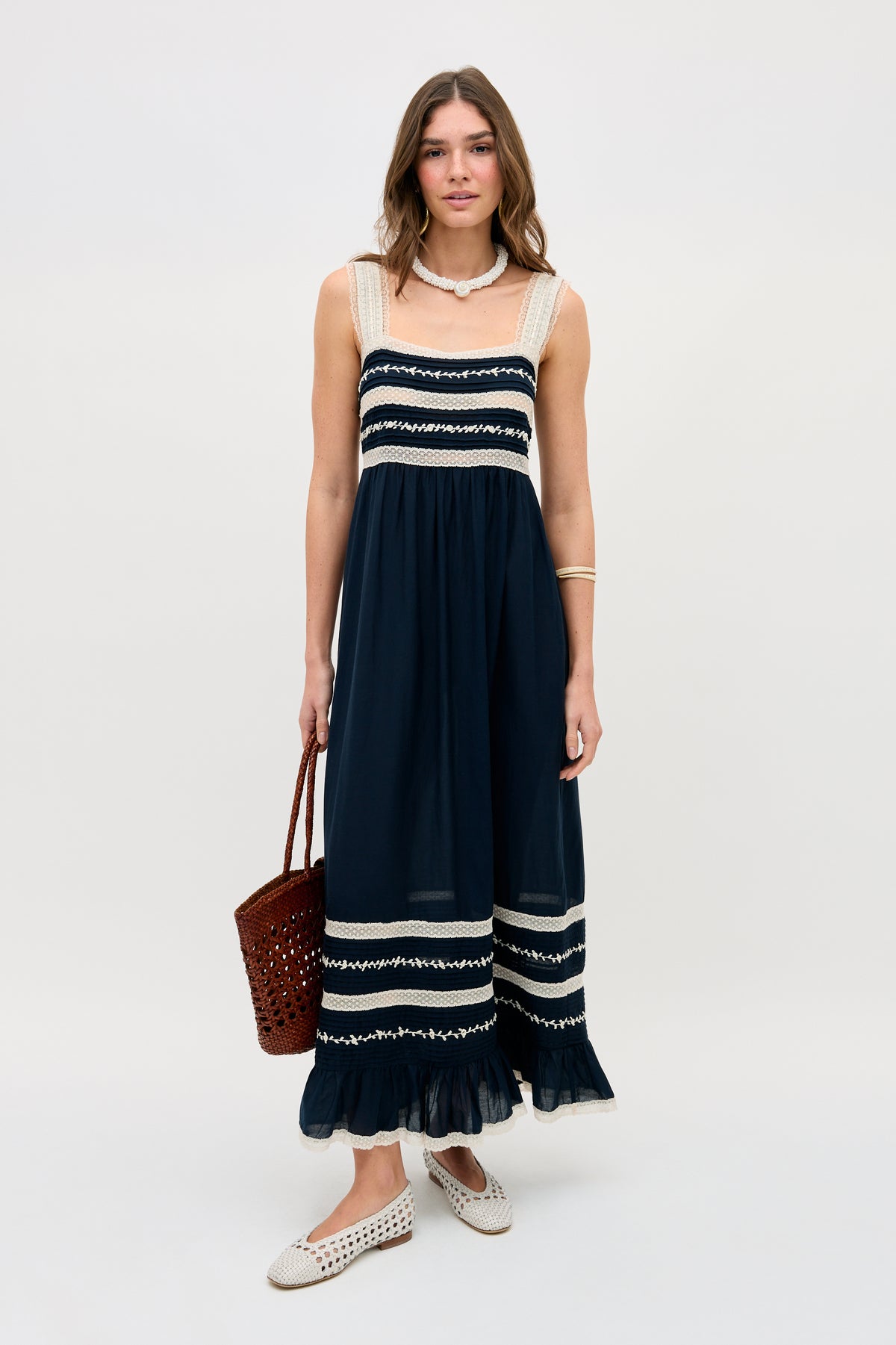 A woman stands against a plain background, wearing the Malie - Deep Navy sleeveless maxi dress with white lace trim, paired with a brown woven handbag, white slip-on shoes, and a necklace.