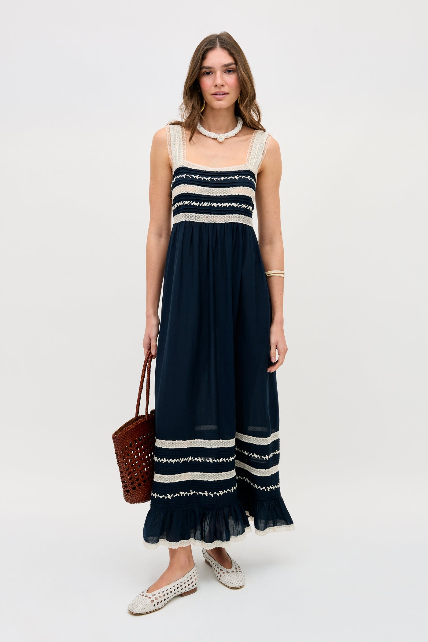 A woman stands against a plain background, wearing the Malie - Deep Navy sleeveless maxi dress with white lace trim, paired with a brown woven handbag, white slip-on shoes, and a necklace.