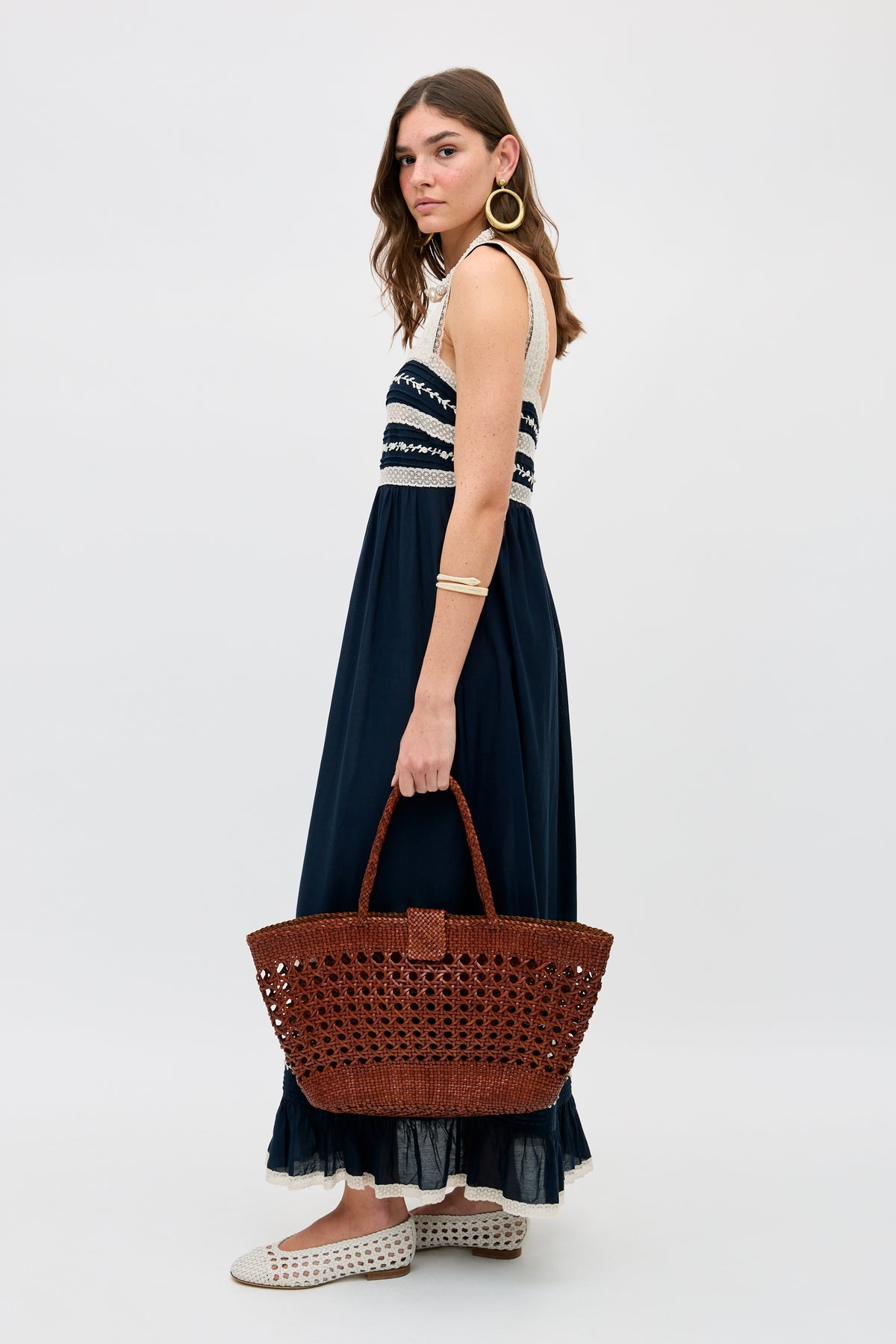 A woman wears the Malie - Deep Navy sleeveless maxi dress, holding a large brown woven tote. She pairs it with white woven slip-on shoes, a gold bracelet, and hoop earrings, standing against a plain light background.