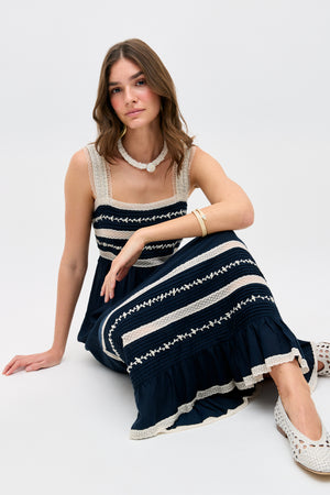 A woman with wavy brown hair wears the Malie - Deep Navy sleeveless embroidered dress. Seated on the floor with one leg bent, she looks calmly at the camera, accessorized with a necklace, bracelet, and white shoes.