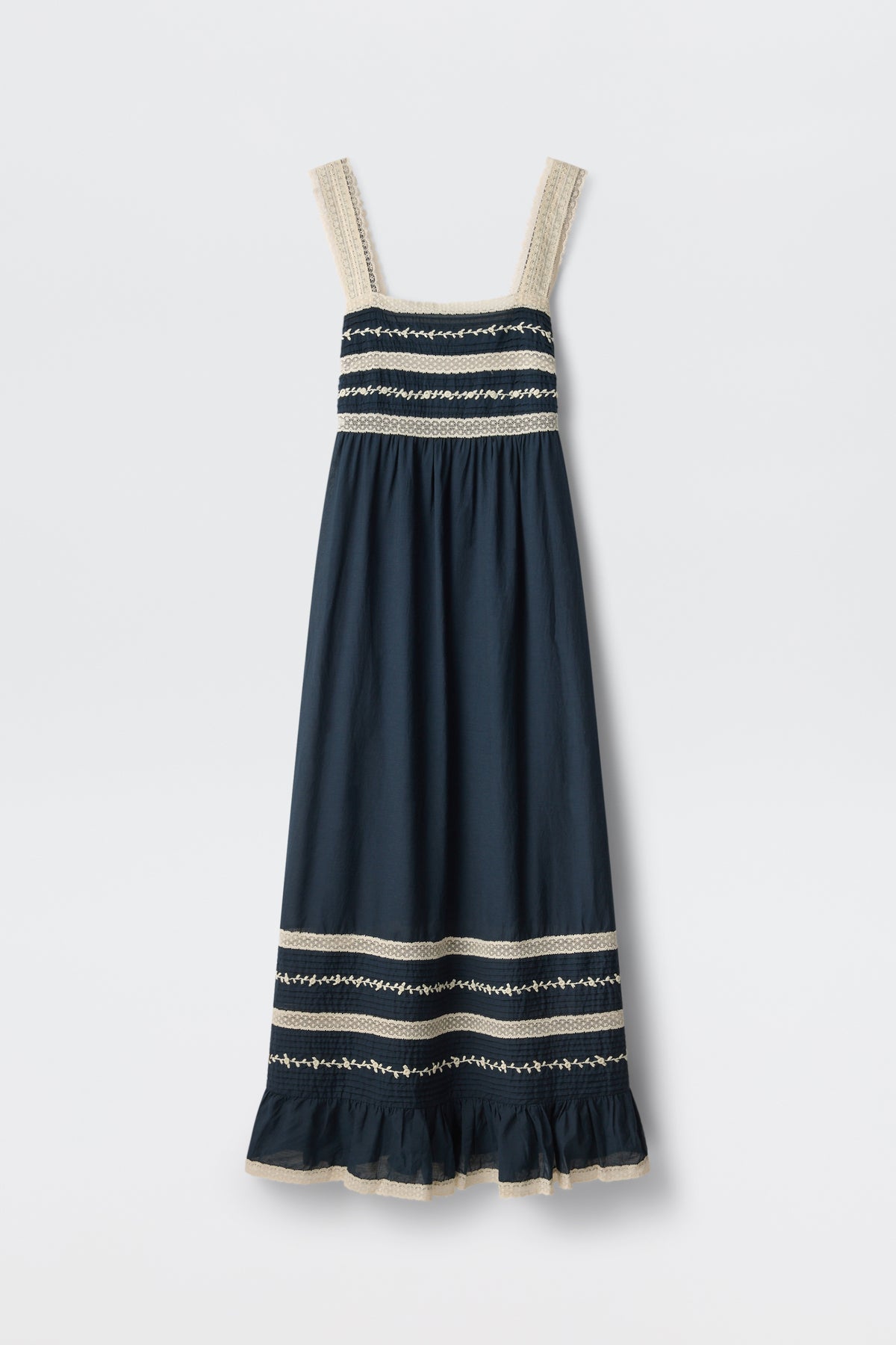The Malie - Deep Navy is a sleeveless maxi dress featuring wide cream lace-trimmed straps, cream and white embroidered stripes on the bodice and hem, and a ruffled bottom edge.