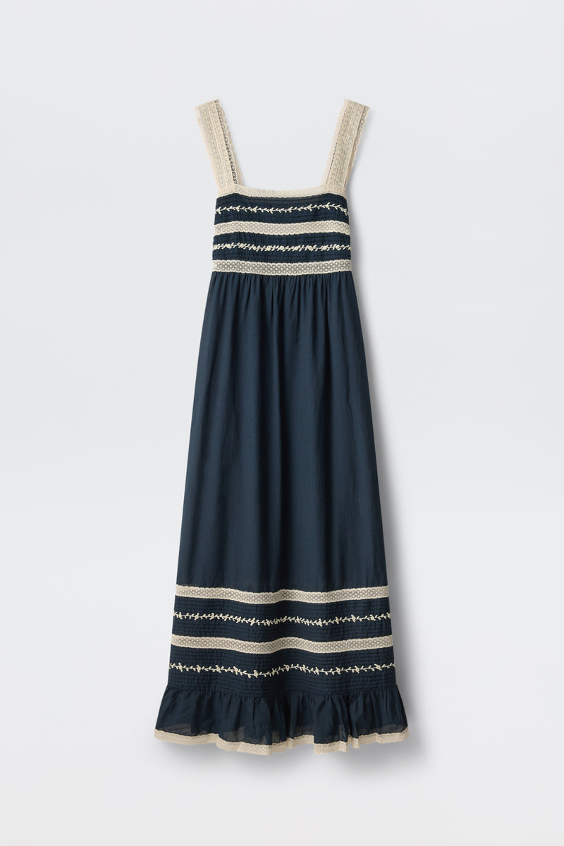The Malie - Deep Navy is a sleeveless maxi dress featuring wide cream lace-trimmed straps, cream and white embroidered stripes on the bodice and hem, and a ruffled bottom edge.