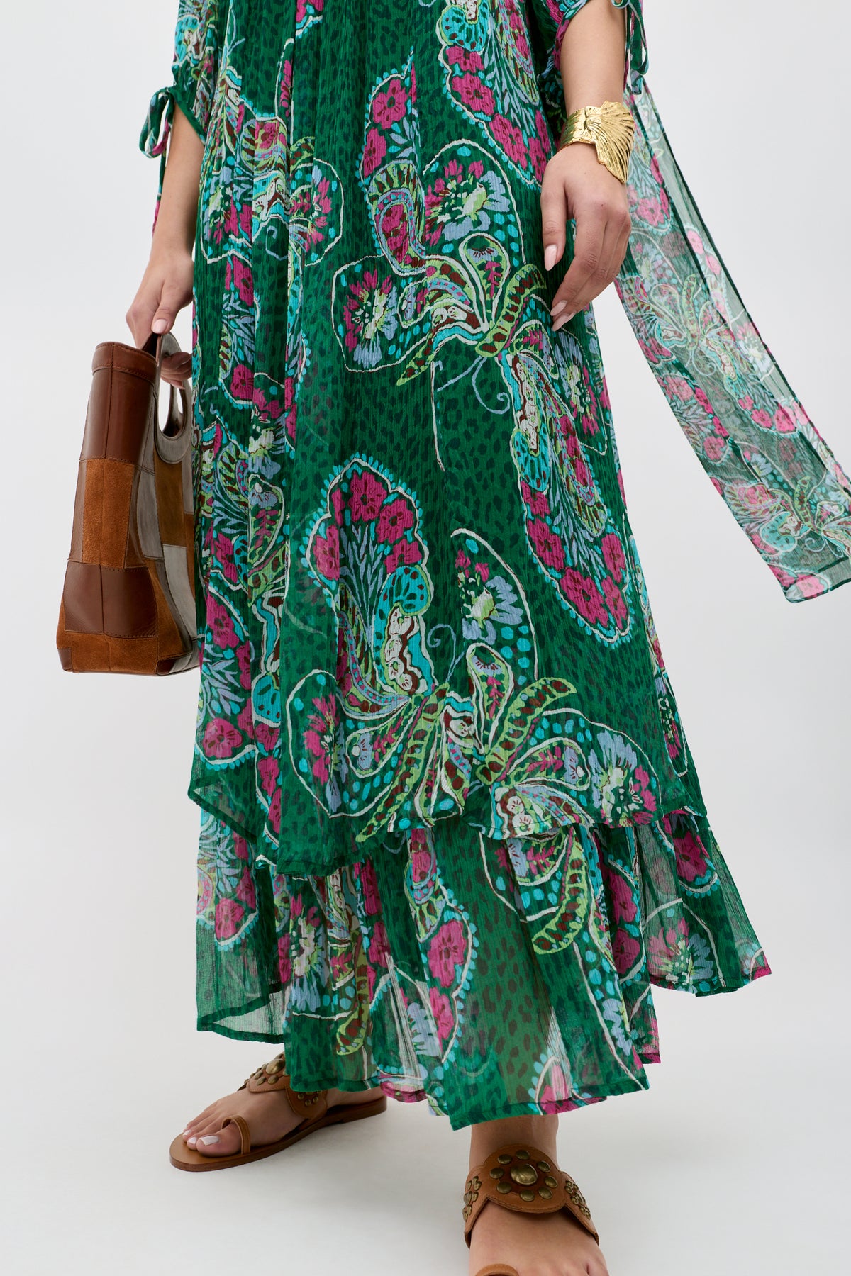 A person wearing the Hayden - Butterfly Bloom Green dress, paired with brown sandals, a gold bracelet, and holding a brown and clear handbag. Only the lower half and one arm are visible.